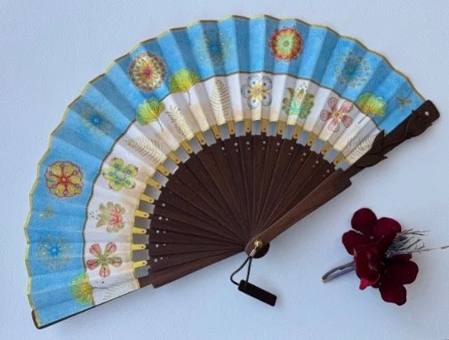 A decorative folding hand fan with floral patterns, blue and white sections, stored on a light surface next to a small bunch of red fabric flowers.