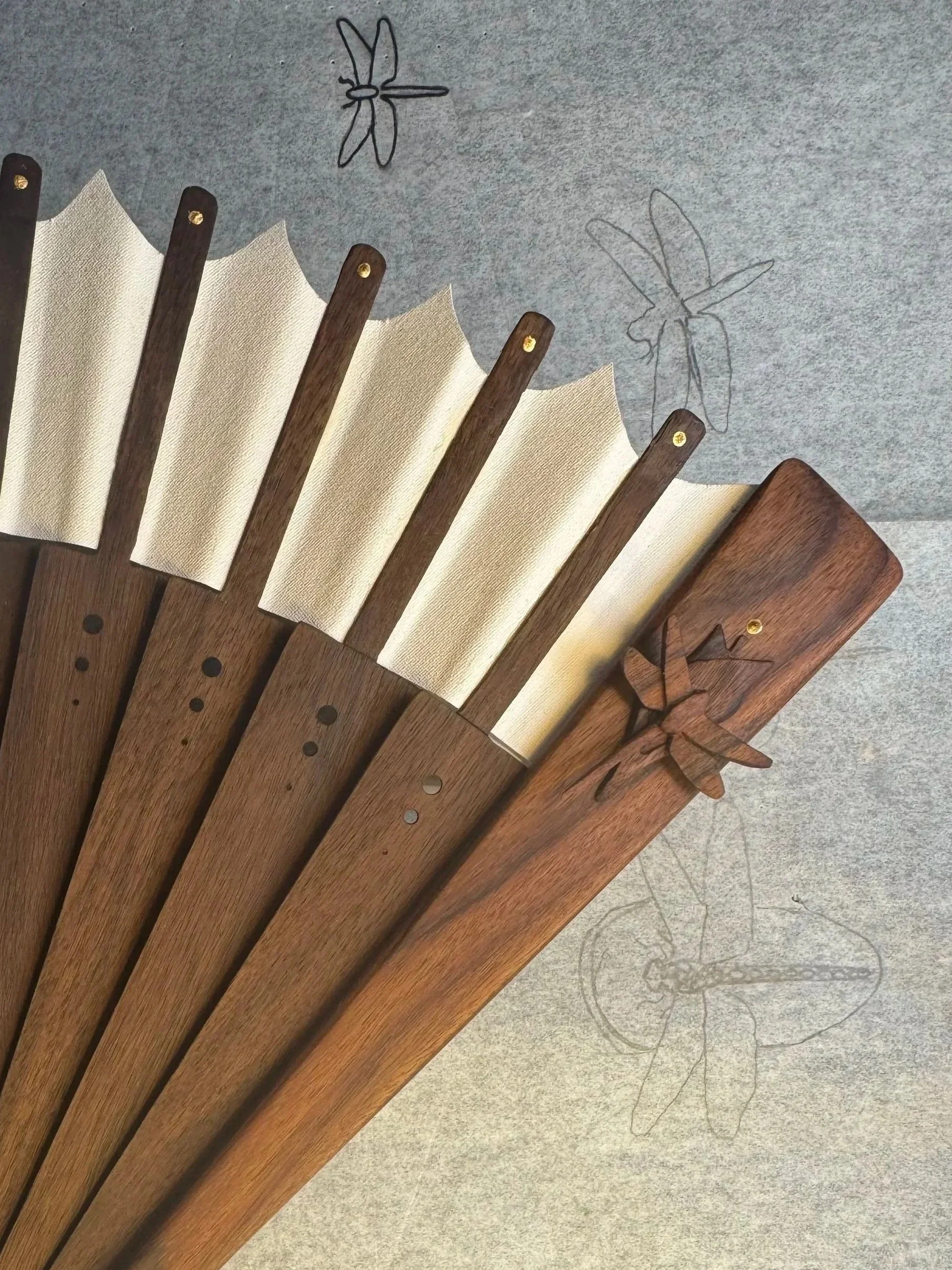 A wooden hand fan with gold accents and beige segments displayed on fabric with dragonfly wood carvings on it.
