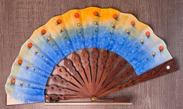 A folding fan with colorful fabric and wooden ribs, decorated with tiny orange and yellow dots and a brown carved wooden handle.