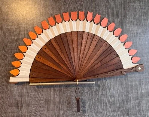 A decorative hand fan with pink and orange paper segments and wooden slats, displayed on a gray surface.