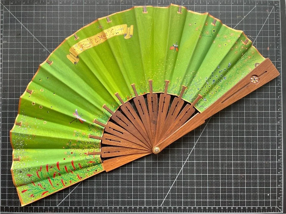 A green decorative folding hand fan with painted dragonflies and fireflies, and a wooden handle, laid open on a black gridded surface.