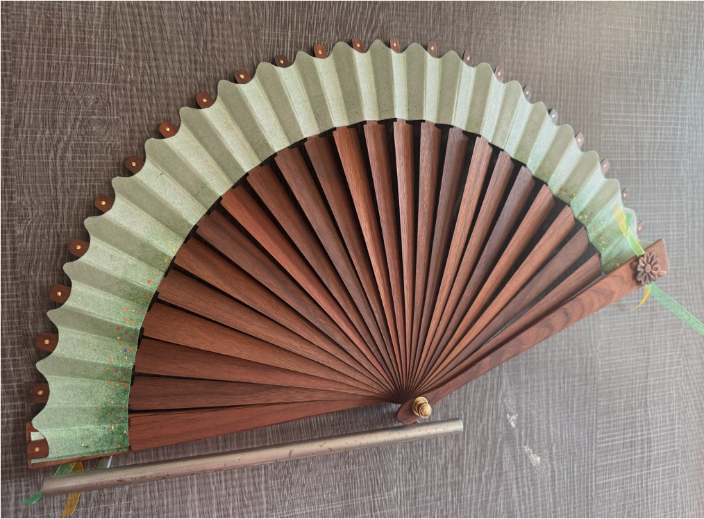 Decorative folding hand fan with wooden ribs, fabric and a floral detail, placed on a gray wooden surface.