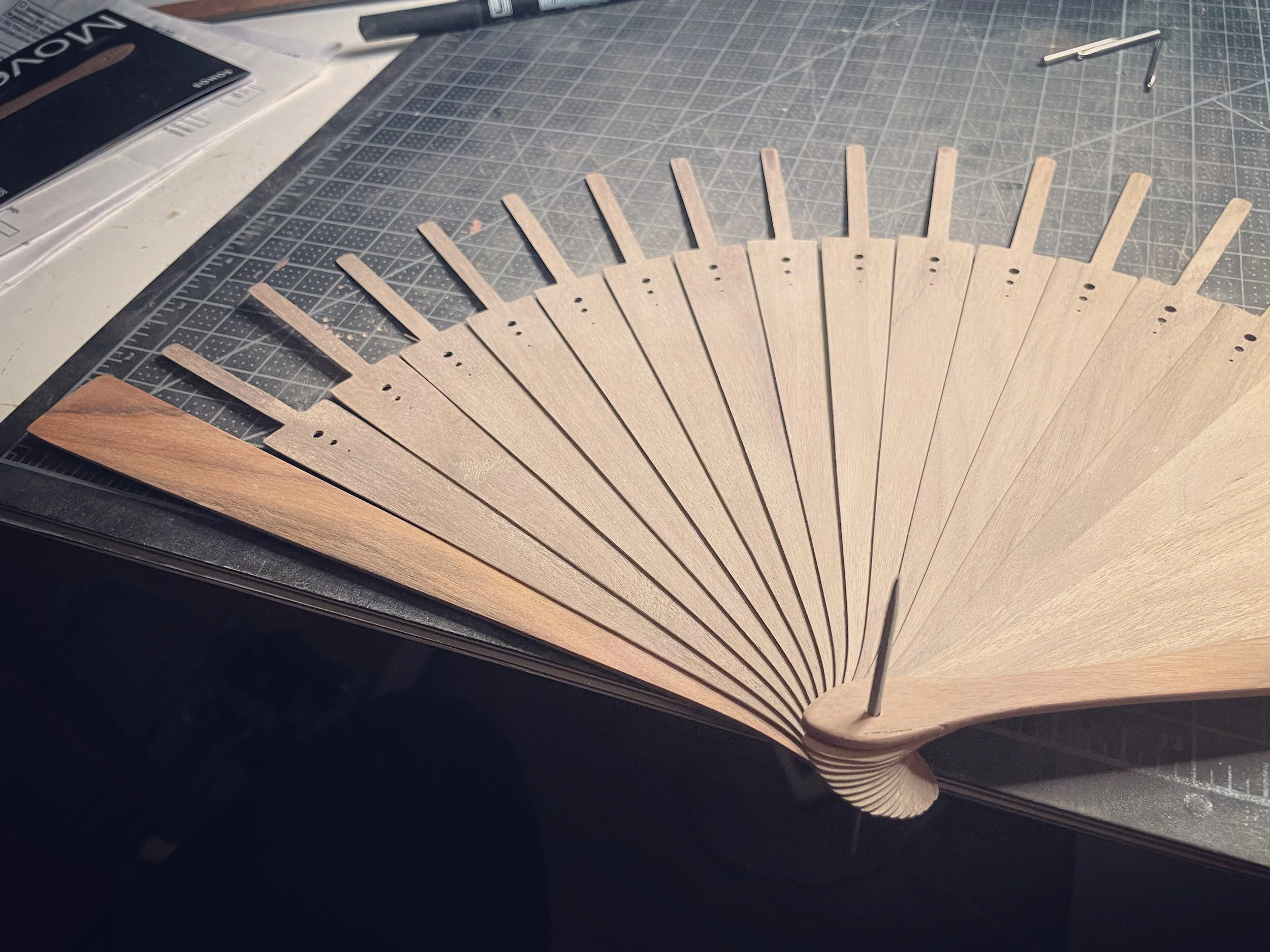 Unfinished wooden hand fan with a pin at the base, laid on a cutting mat with grid lines, with carving tools nearby.