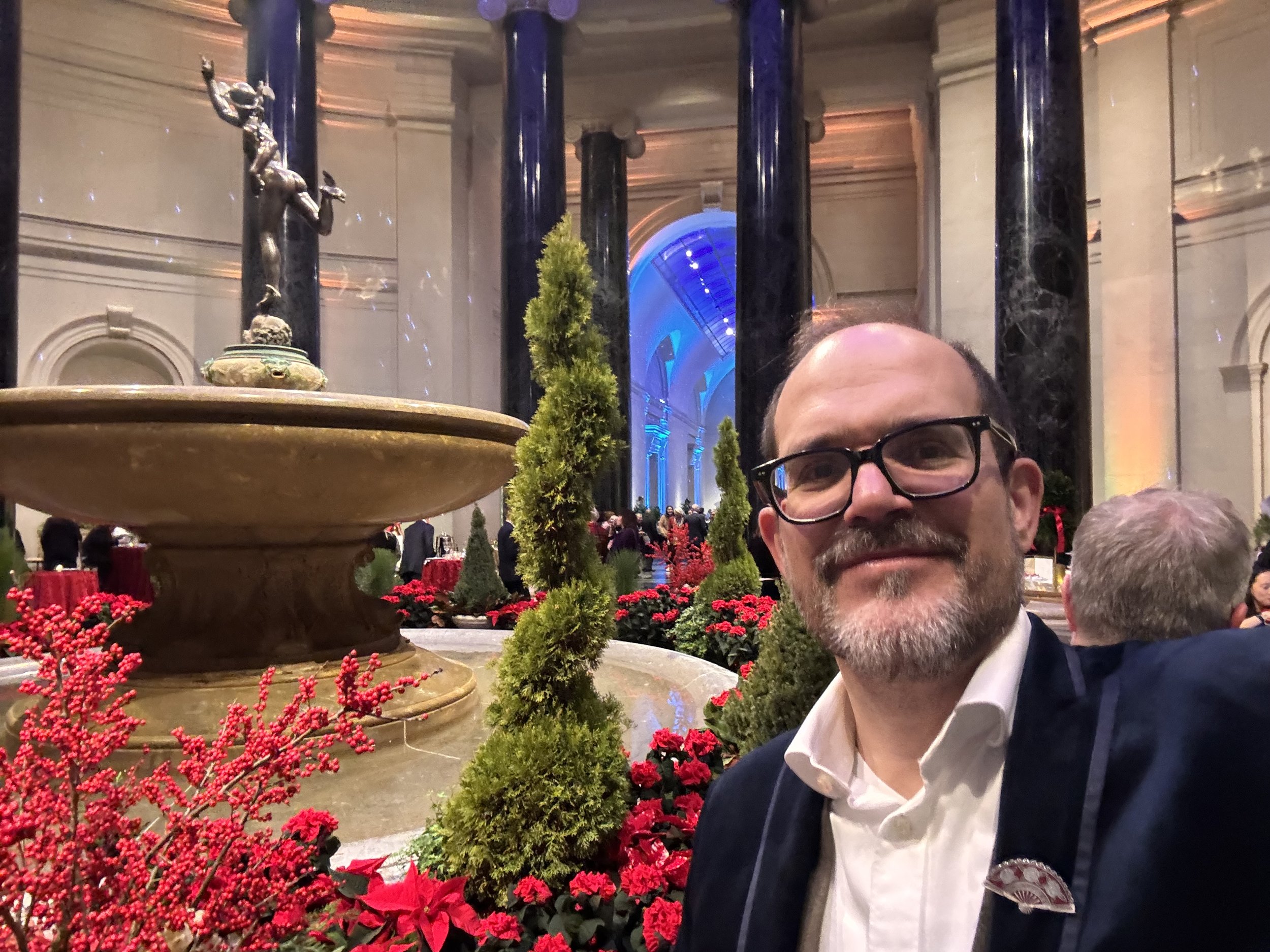 A man with glasses and a beard smiling in front of a decorative fountain at an indoor event with holiday decorations, including red poinsettias and green pine trees, in a grand hall with tall columns and blue lighting.