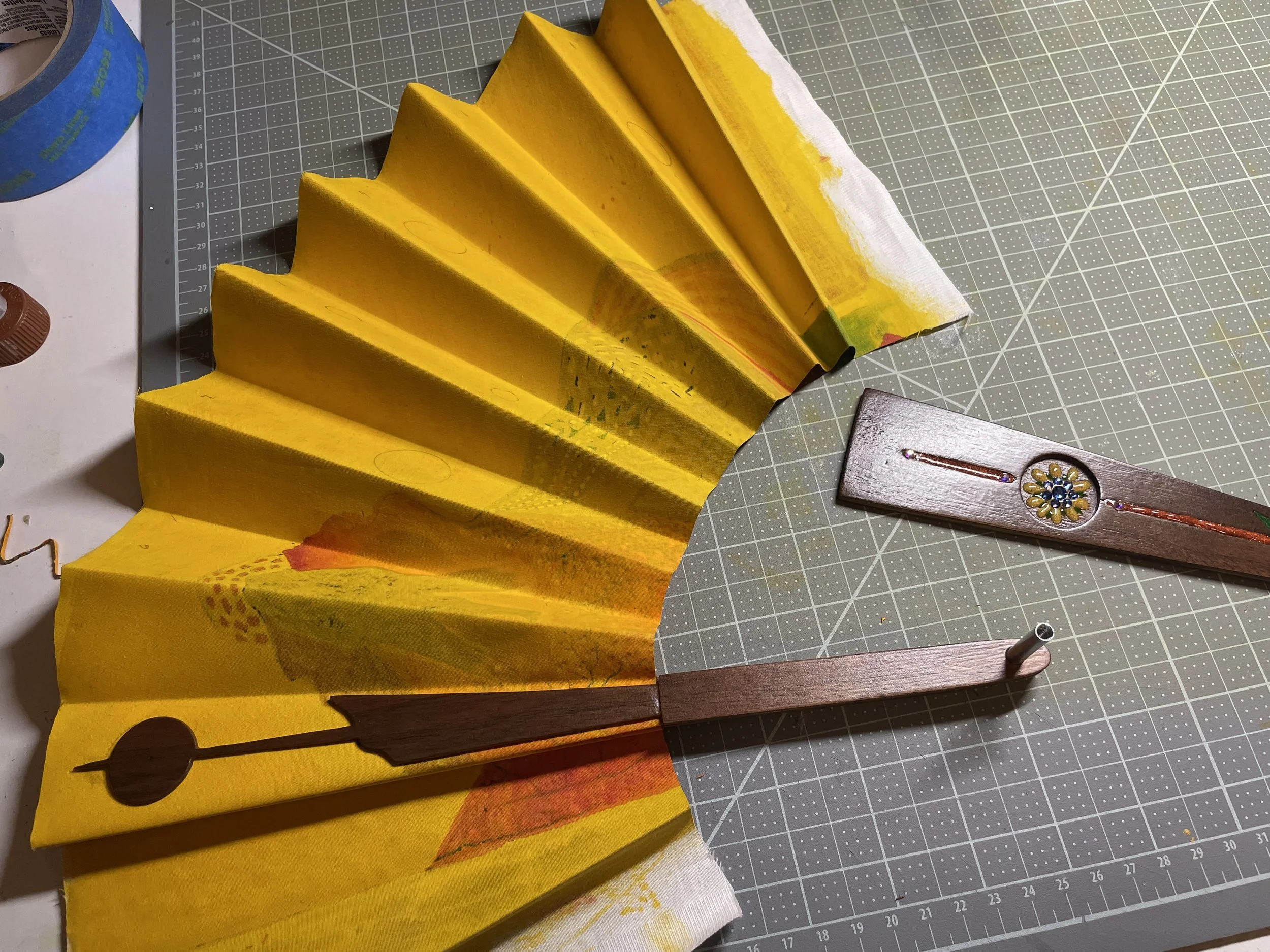 A partially assembled decorative fabric fan with yellow and orange colors, on a crafting grid cutting mat, with paint and craft tools nearby.