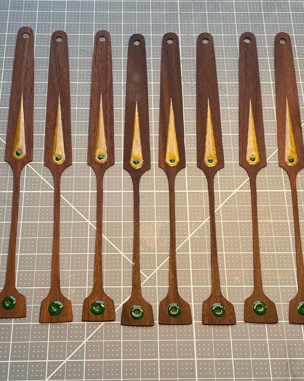 Seven handcrafted wooden fan staves with painted yellow and green accents, including resin inlays, laid out on a grey cutting mat with white grid lines.
