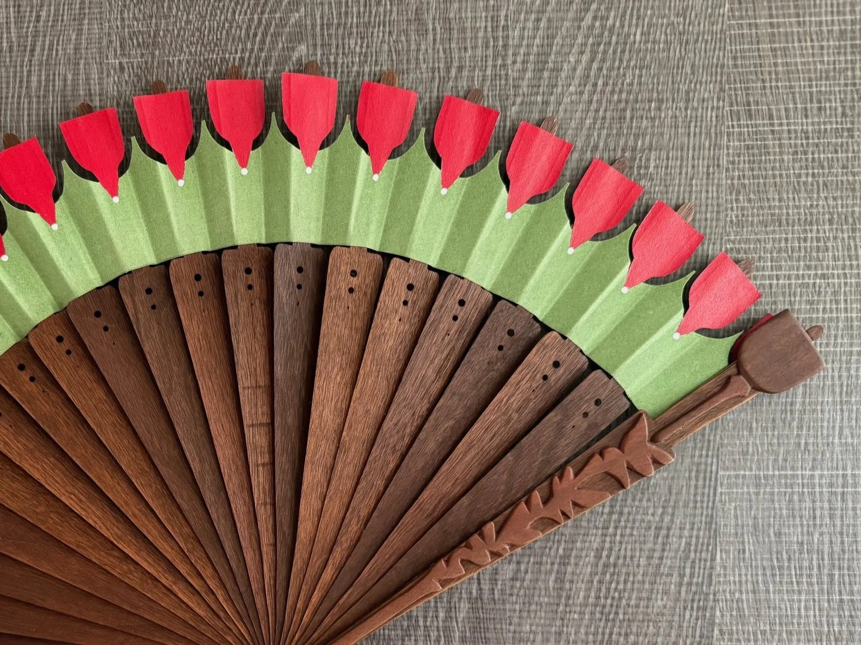 Decorative handheld folding fan with wooden slats and red, pink, and green painted fa bric, with embellishments resembling flowers or petals.