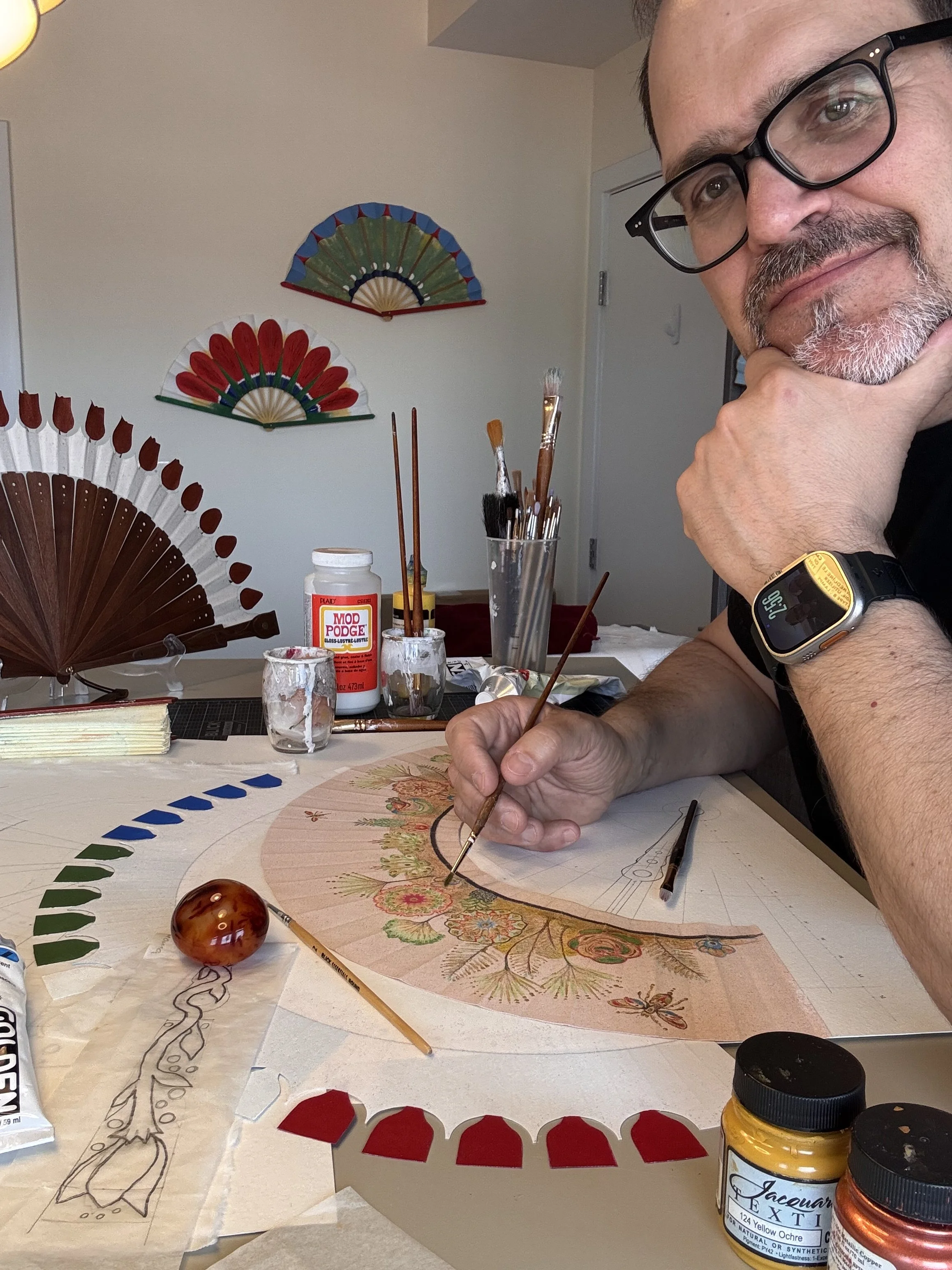 A man with glasses and a beard smiling while working on a painted fan at a table filled with brushes, paint jars, and markers, with decorative fans hanging on the wall in the background.