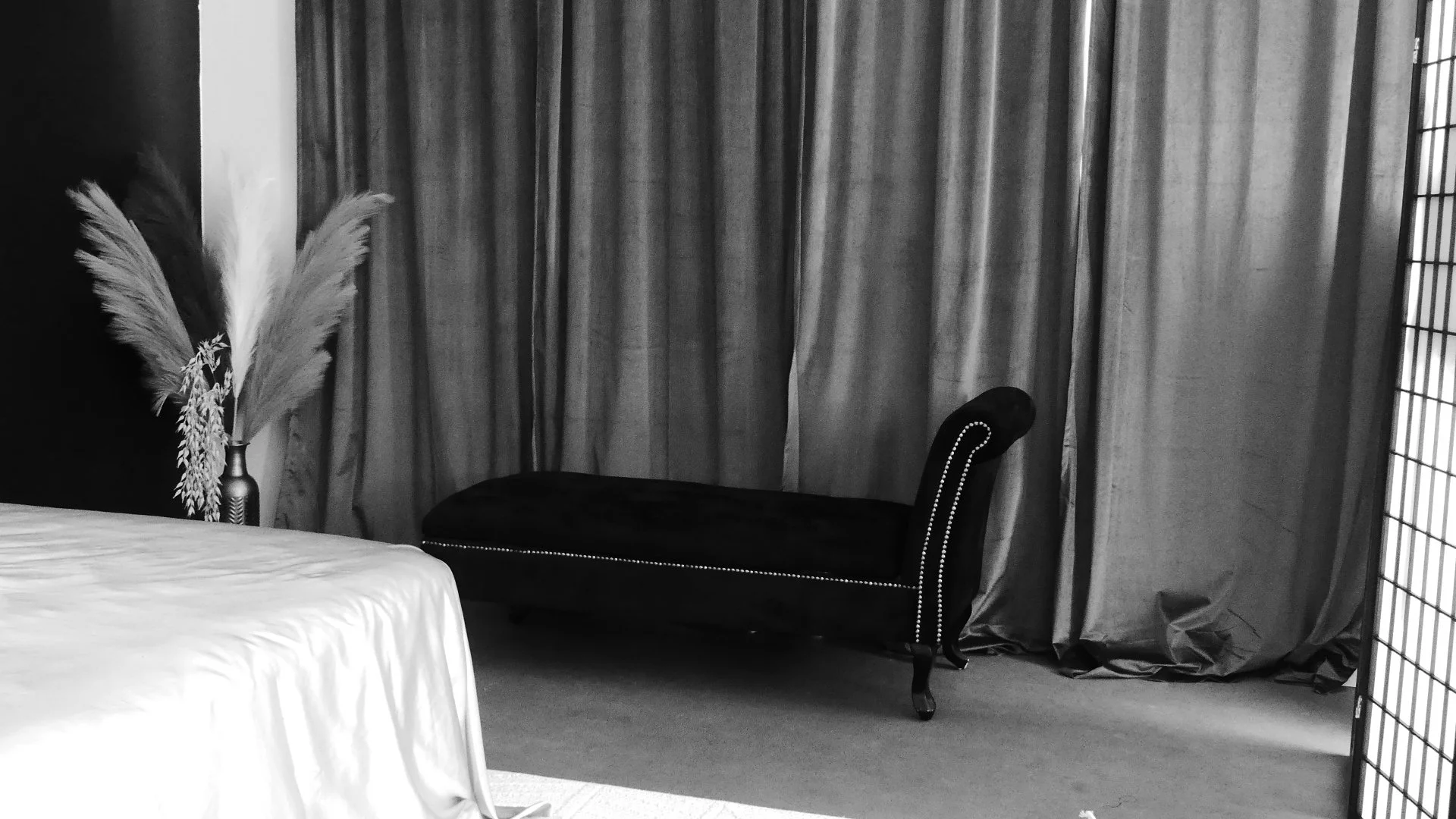 A black velvet chaise lounge with nailhead trim in front of curtains, next to a tall vase with pampas grass on a side table, in a room with a folding screen.