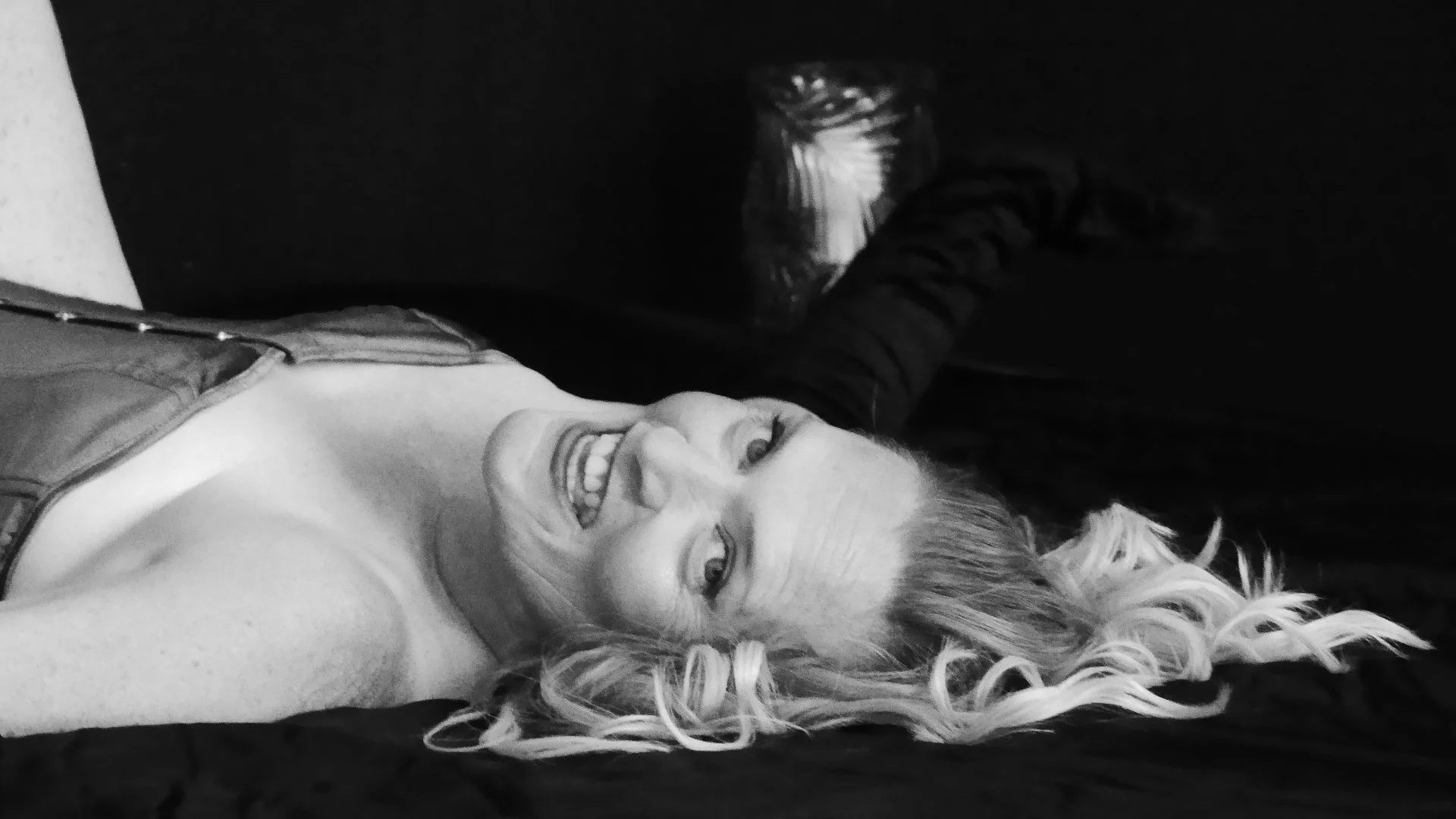A woman with light curly hair lying on their back on a black surface, smiling and looking up, with a glass jar nearby.