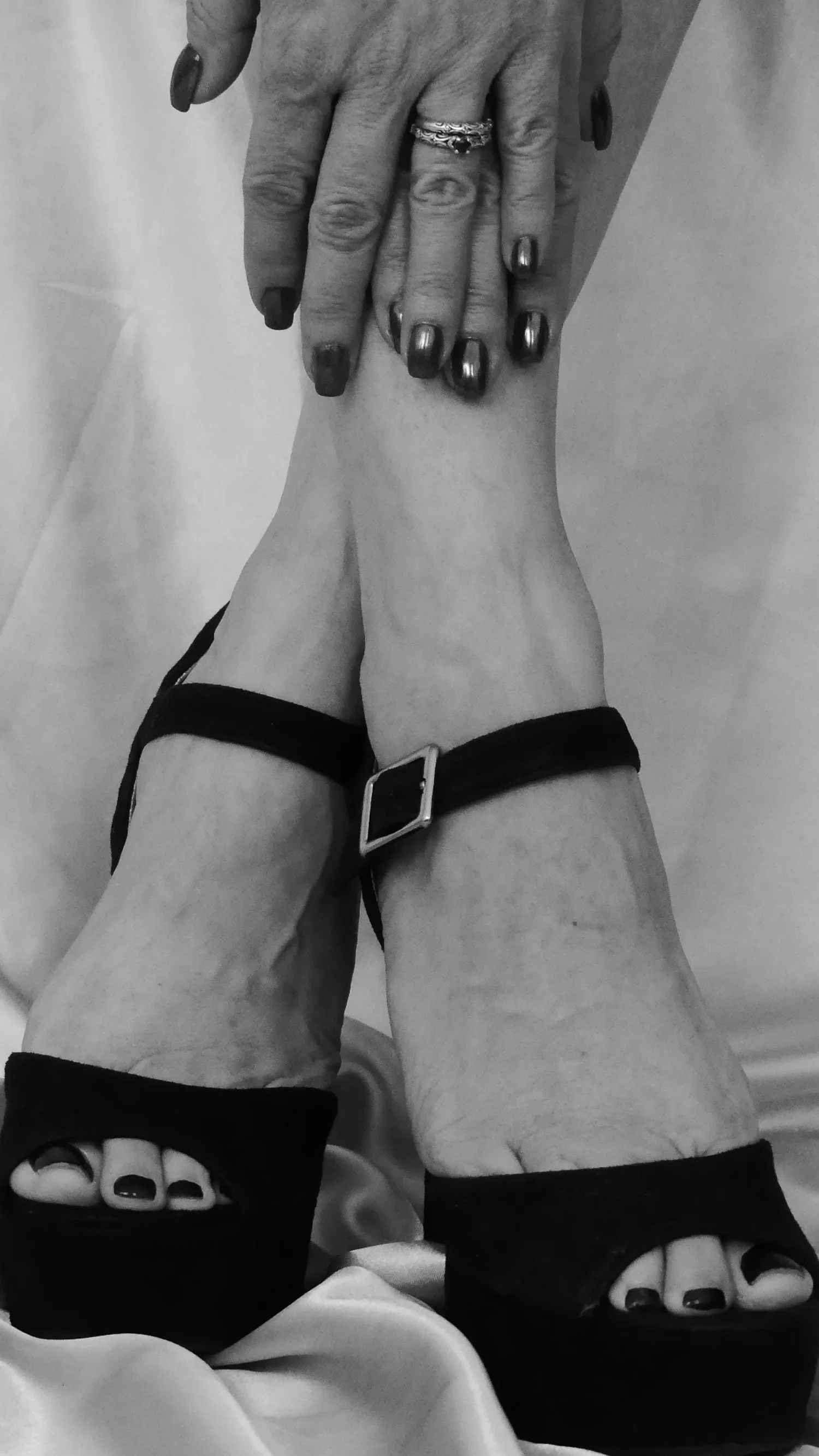 Black and white photo of a woman's hands with rings resting on her ankle, showing her feet in black open-toe heels with painted toenails, resting on satin fabric.