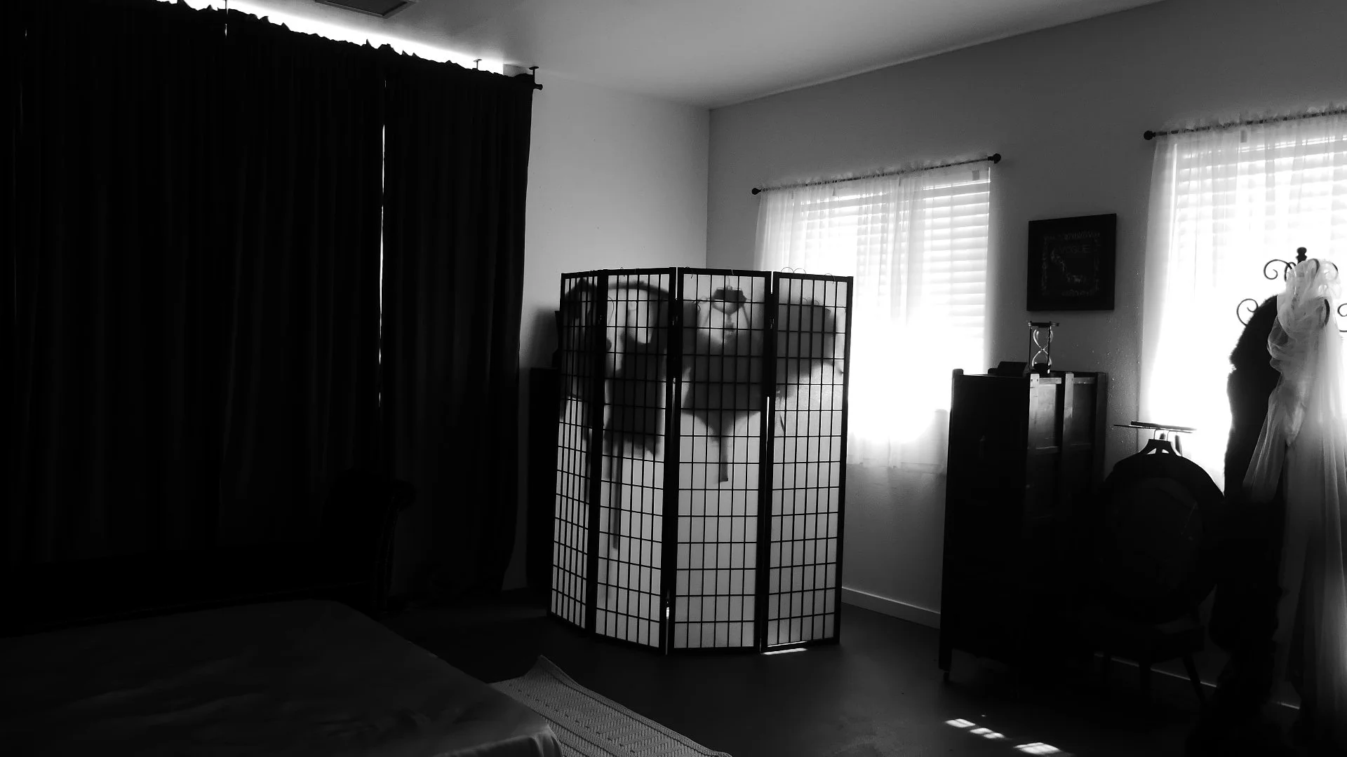 A black and white photo of a bedroom with two windows, one mostly covered by dark curtains and the other with sheer curtains, allowing light in. In the room, there is a folding room divider with clothes hanging on it, a small dresser, and a bridal go