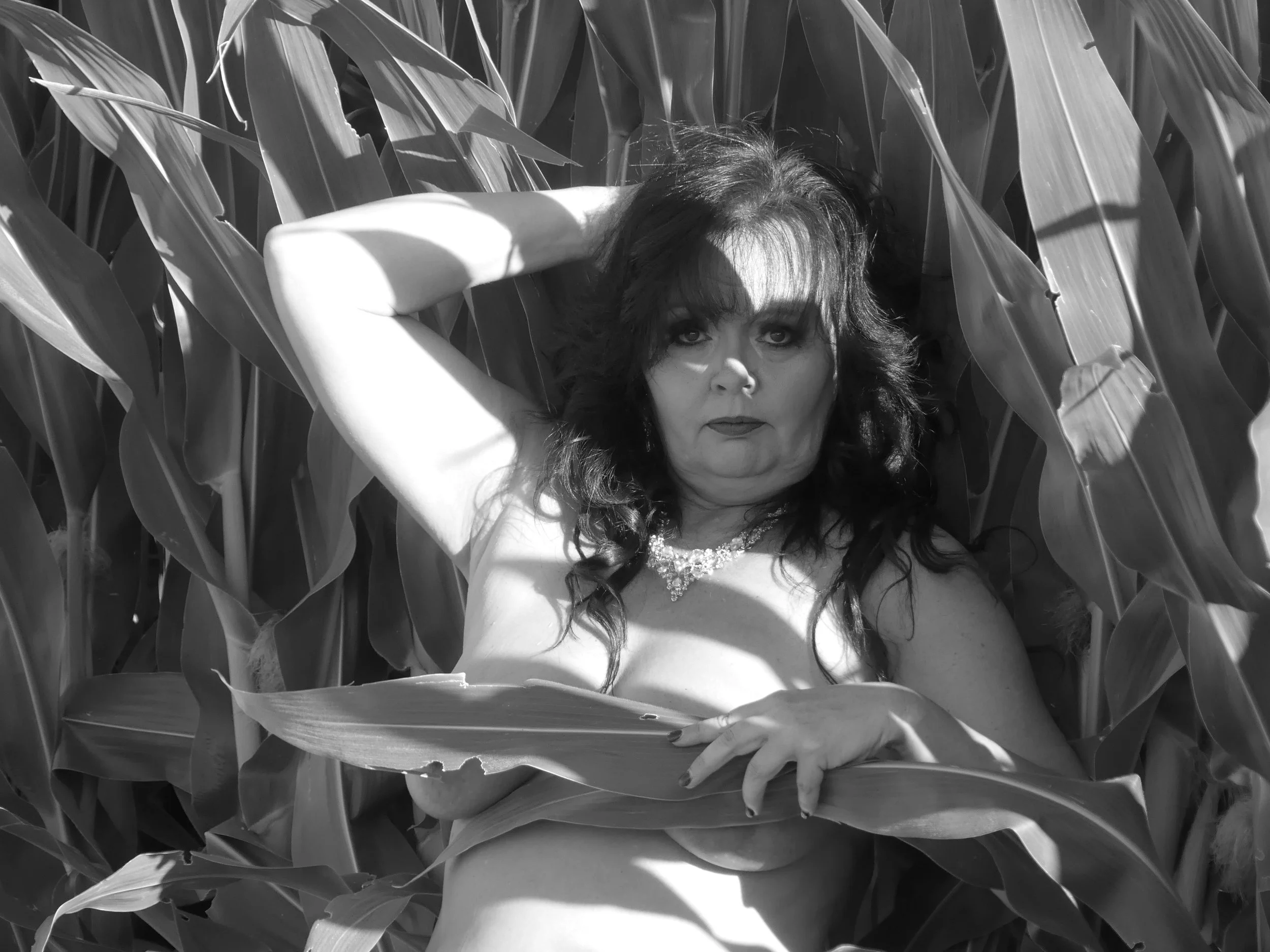 A woman with dark hair and makeup, wearing a necklace, lying among tall corn plants, topless, with one arm above her head and the other hand holding a leaf.