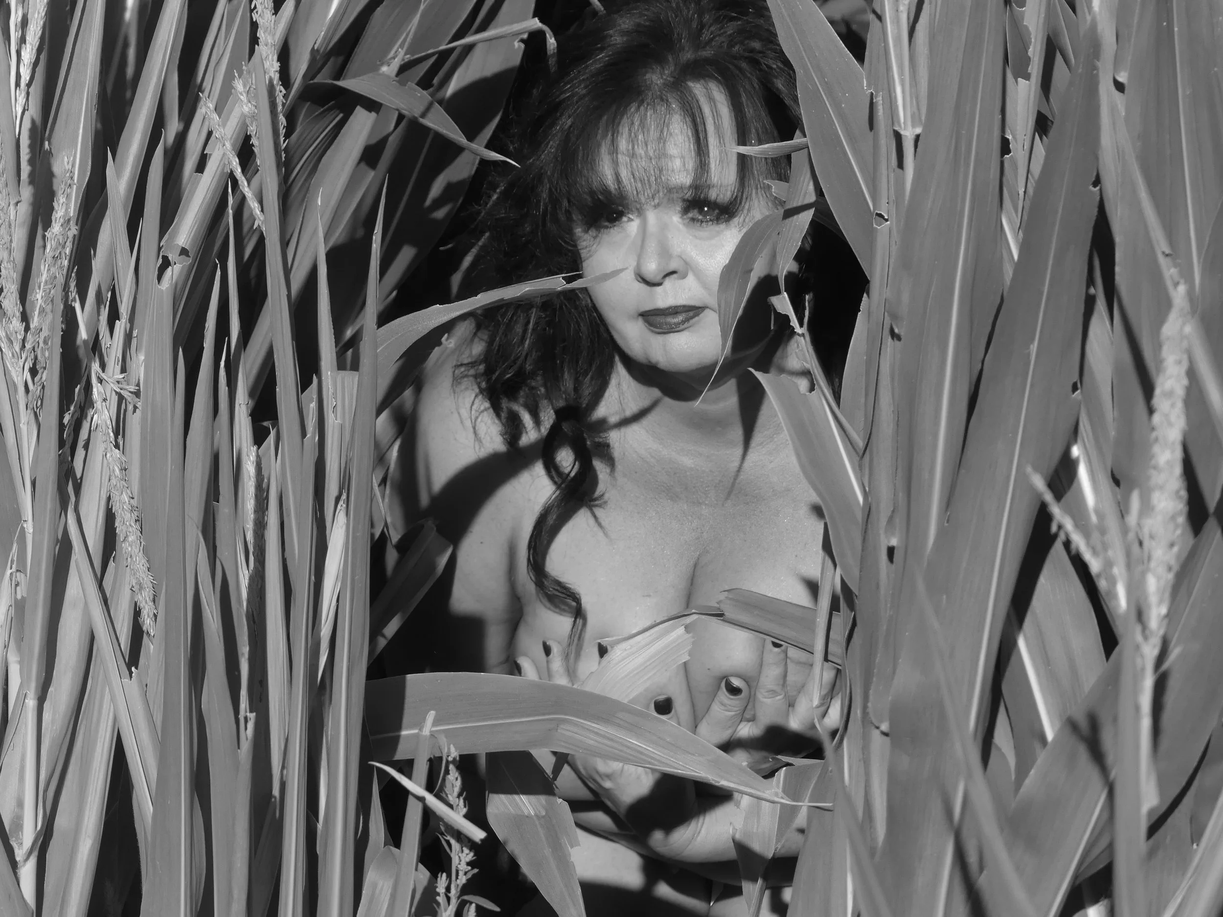 A woman with dark, wavy hair and makeup, partially nude, is crouching among tall, leafy plants. Her face and upper body are visible, with her hand on her chest, framed by the leaves.
