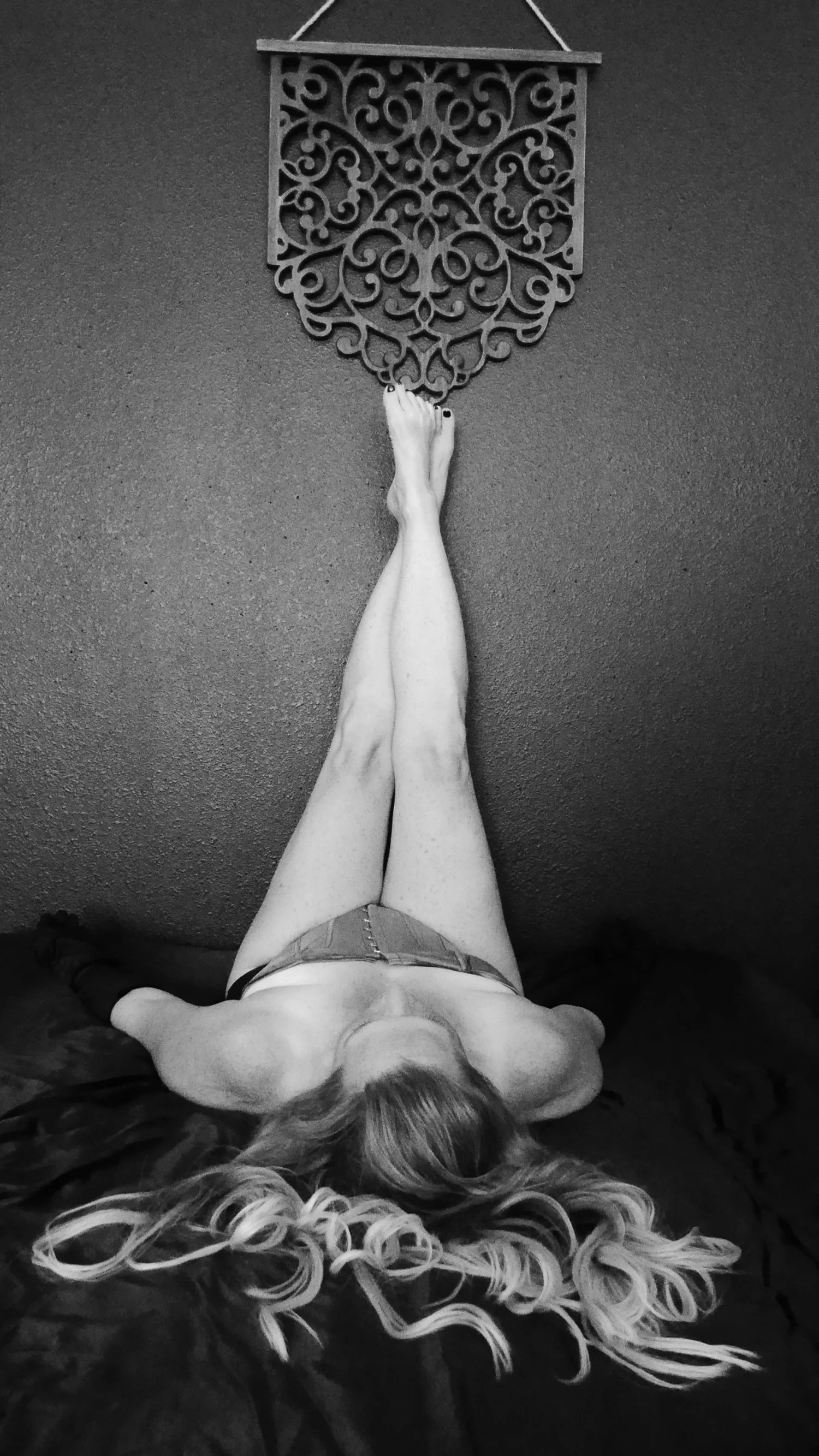 A black and white photo of a woman with long wavy hair lying on her back on a bed, with her legs extended upward against a wall. She is wearing shorts and a top, and is touching a decorative wall hanging above her feet.