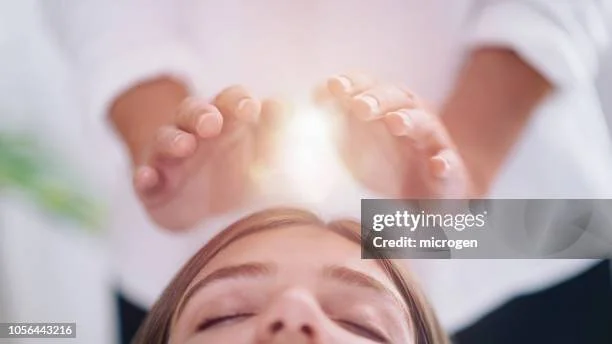 A person lying down with eyes closed receiving a healing or energy treatment from another person using their hands positioned above the head, emitting a radiant glow.