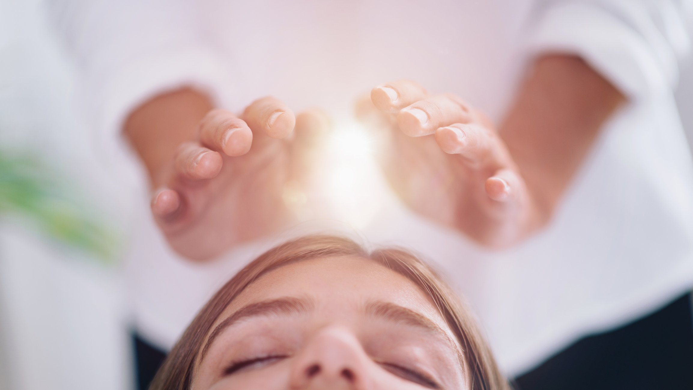 A person lying down with eyes closed receiving a healing or energy treatment from another person using their hands positioned above the head, emitting a radiant glow.