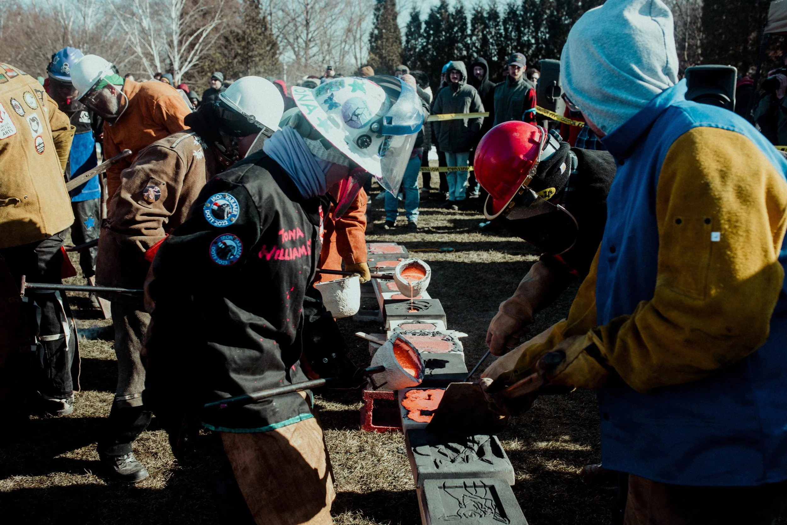 Workers pouring molten metal into molds at an outdoor event, with onlookers in the background.
