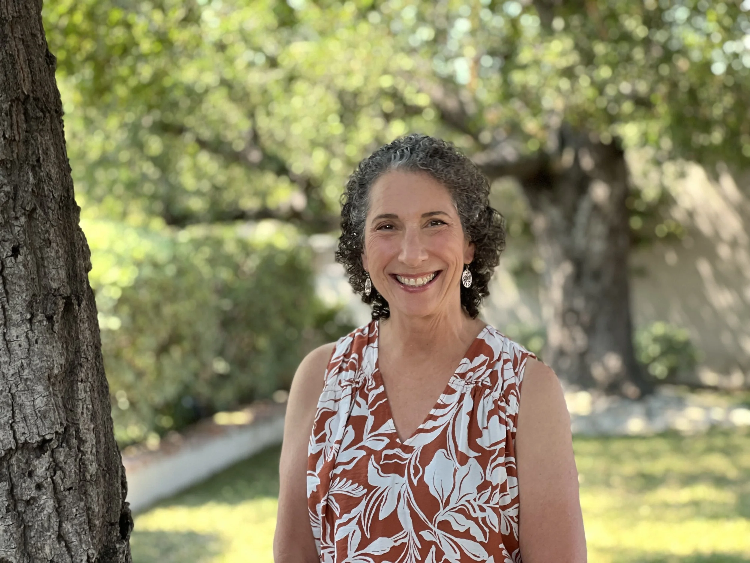 Sue Brian, a woman with curly gray hair smiling outdoors in a garden, wearing a sleeveless dress with a white and brown floral pattern and silver earrings.