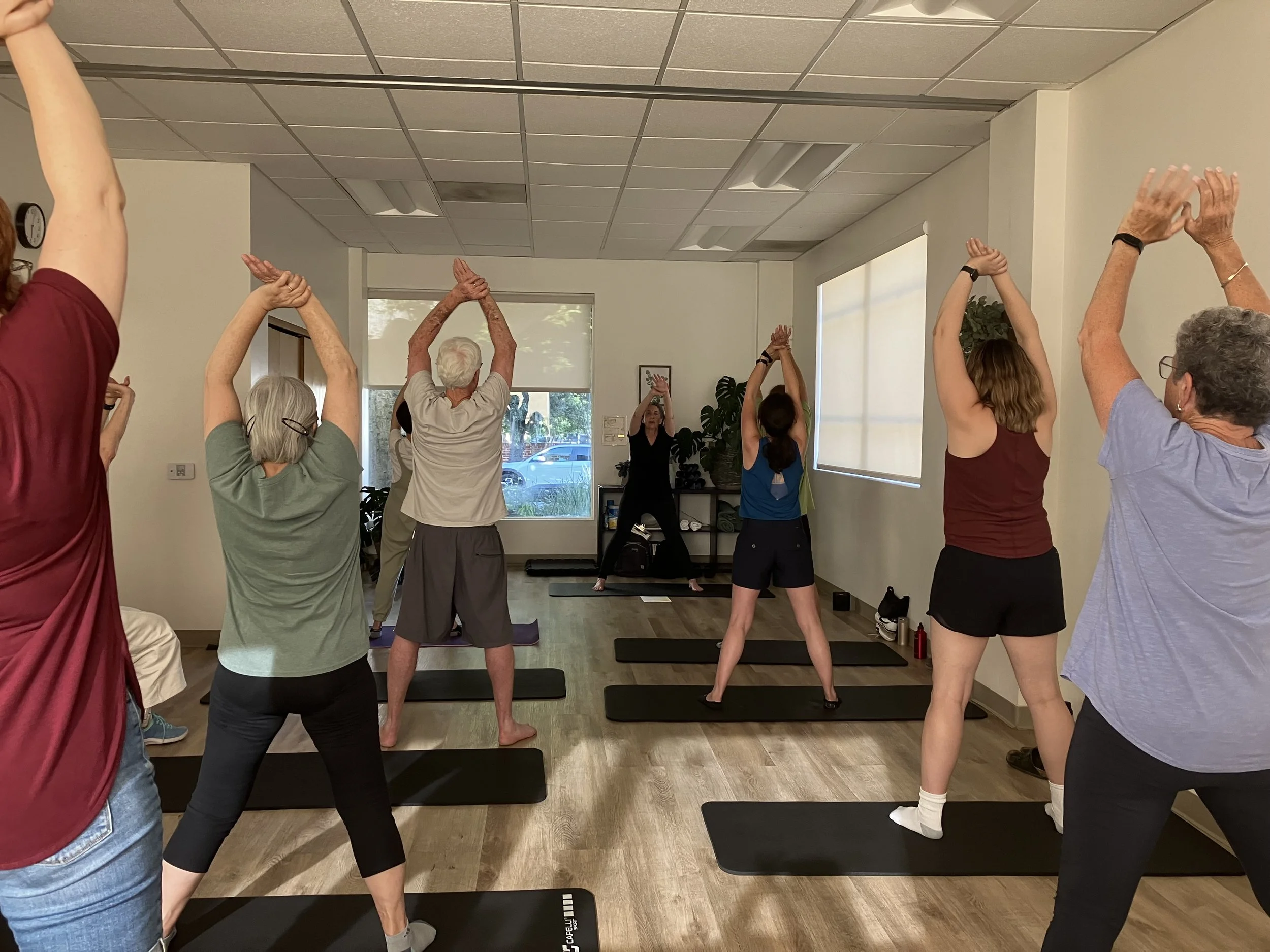 People participating in a yoga class in a room with large windows, stretching above their heads on yoga mats. Photo taken from attending an in person class with Sue Brian