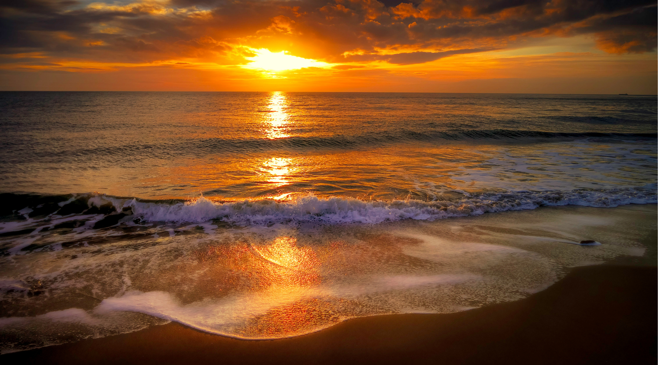 Sunset over the ocean with waves gently crashing on the sandy beach, reflecting the vibrant colors of the sky.