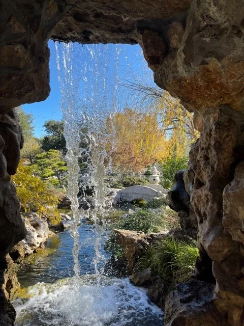View of a garden with a small waterfall through a rocky opening, trees, water, and a stone arch bridge in the background.  Image provided by Cyndi Bemel (cyndibemel.com | cbemel3@me.com)