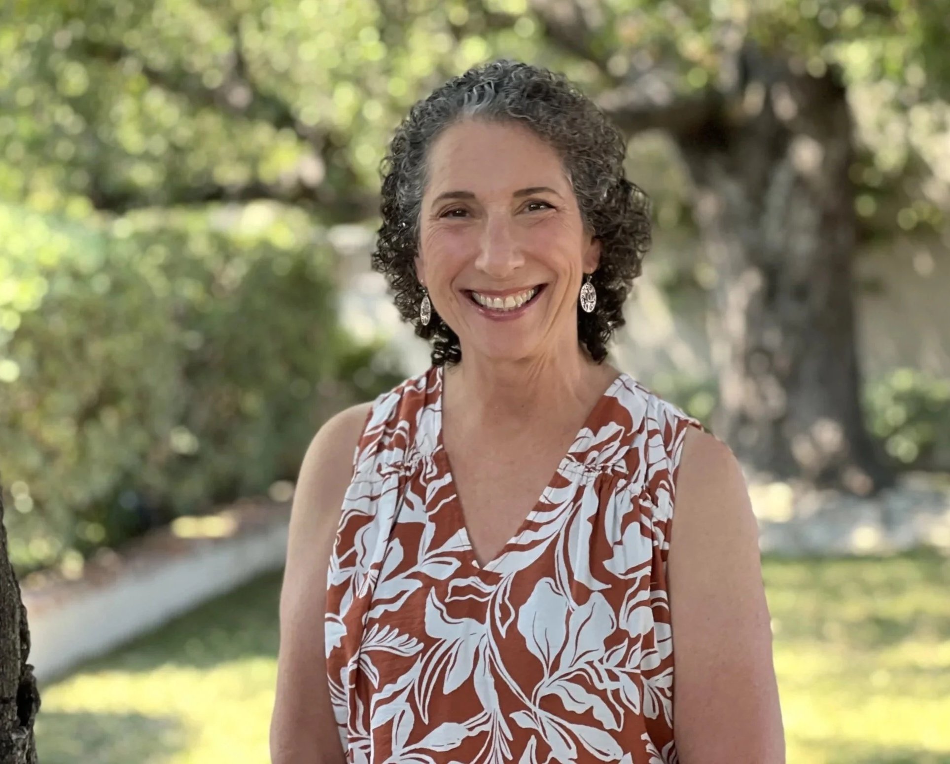 Sue Brian, founder of Two Oaks Method and instructor for , a woman with short curly gray hair smiling outdoors, wearing a sleeveless patterned top and dangle earrings, with a background of trees and greenery.