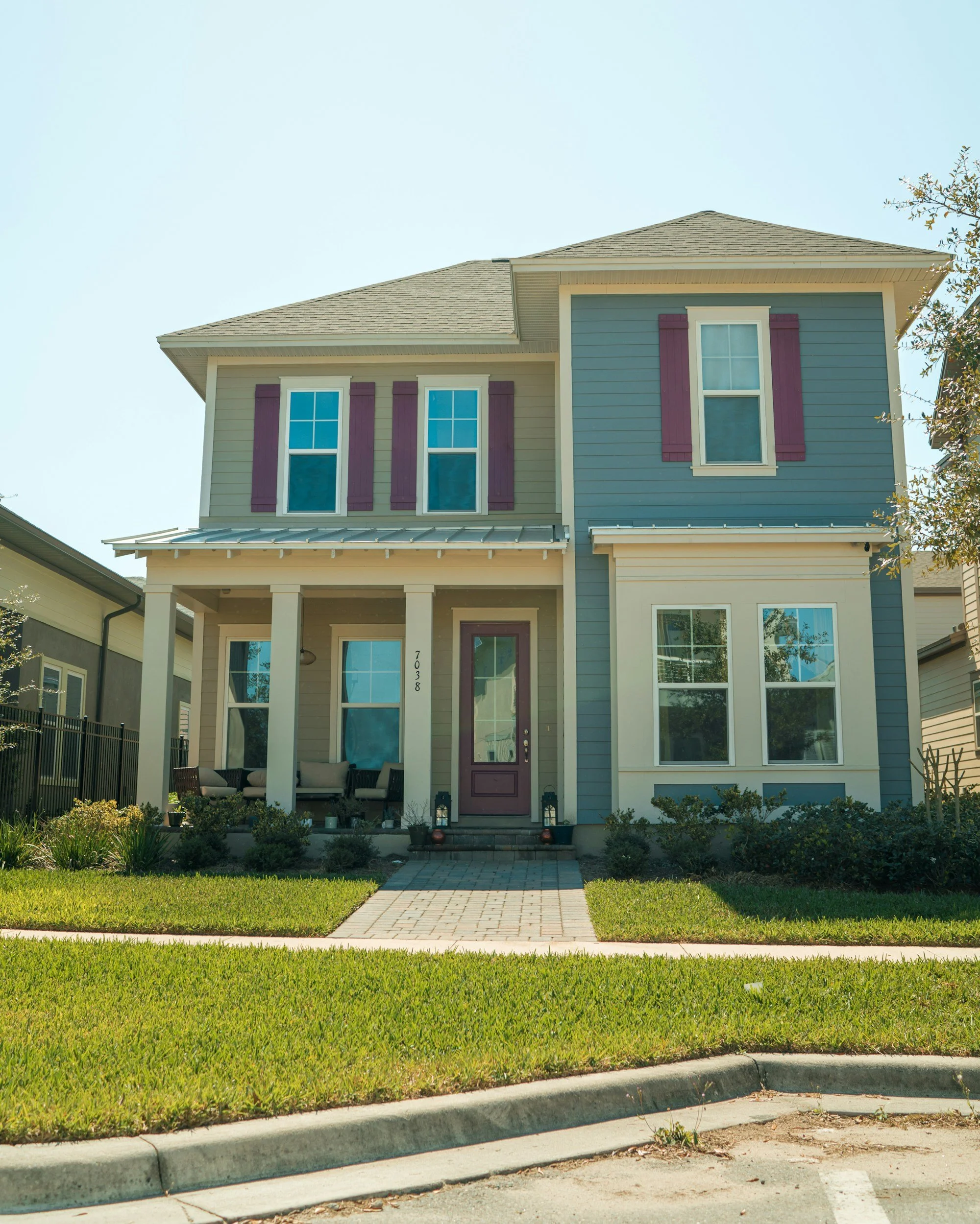 A two-story house with blue and beige siding, purple window shutters, a front porch with seating, a brick pathway, and a well-maintained lawn.