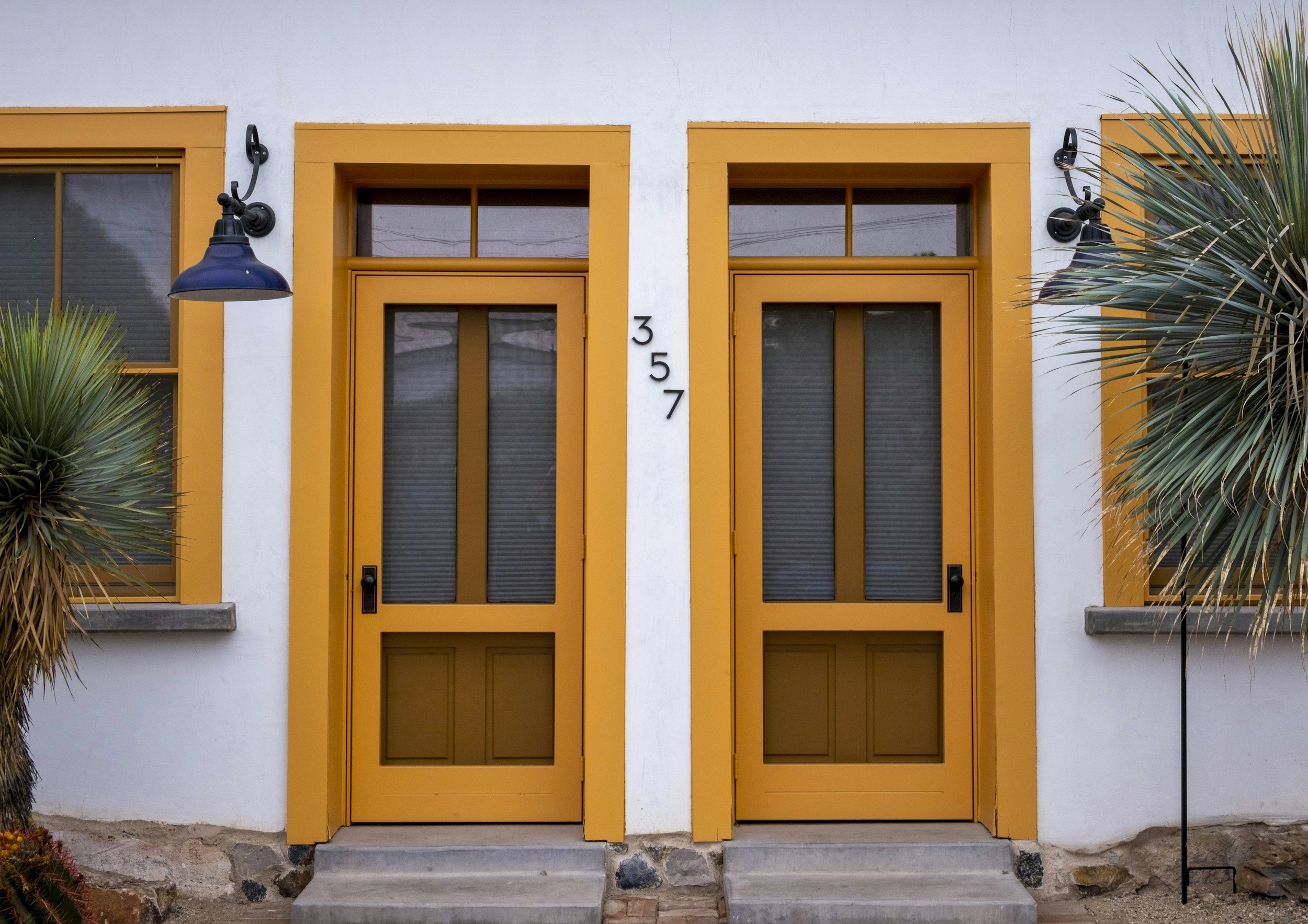 Two yellow front doors with black handles and transom windows, set in a white building with the number 357 vertically between them, flanked by two black outdoor wall lanterns and two large expressive palm trees.