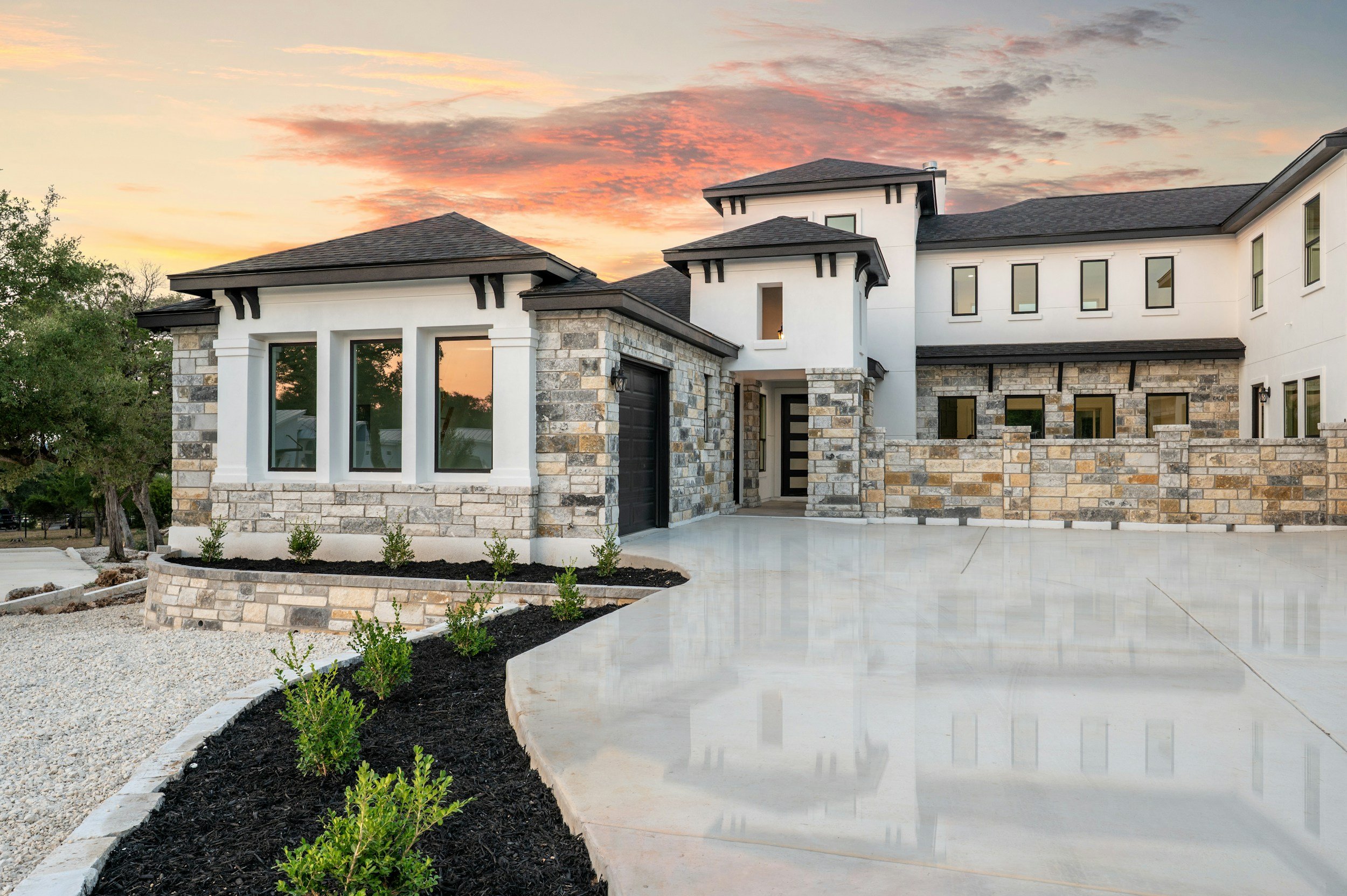 Modern house with stone and white exterior, large windows, black roof, paved driveway, landscaped front yard, and sunset sky in the background.
