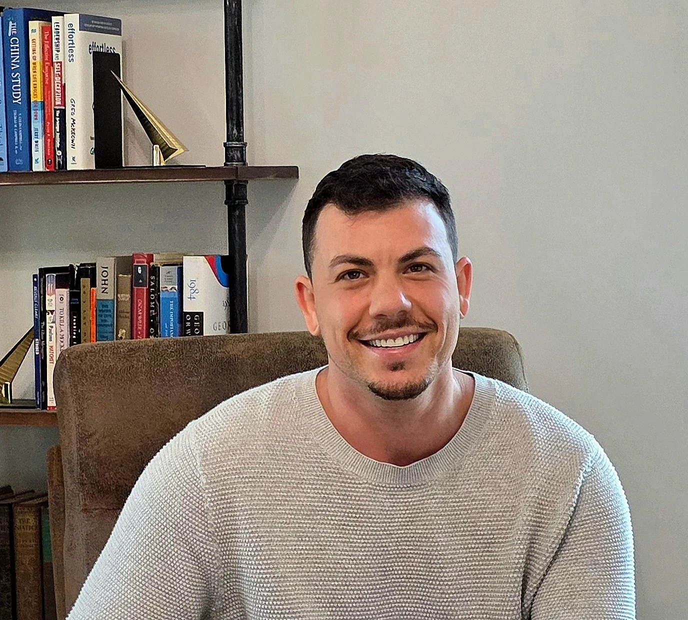 A smiling man with short dark hair, facial hair, wearing a light gray sweater, sitting in a brown armchair with a bookshelf filled with books behind him.