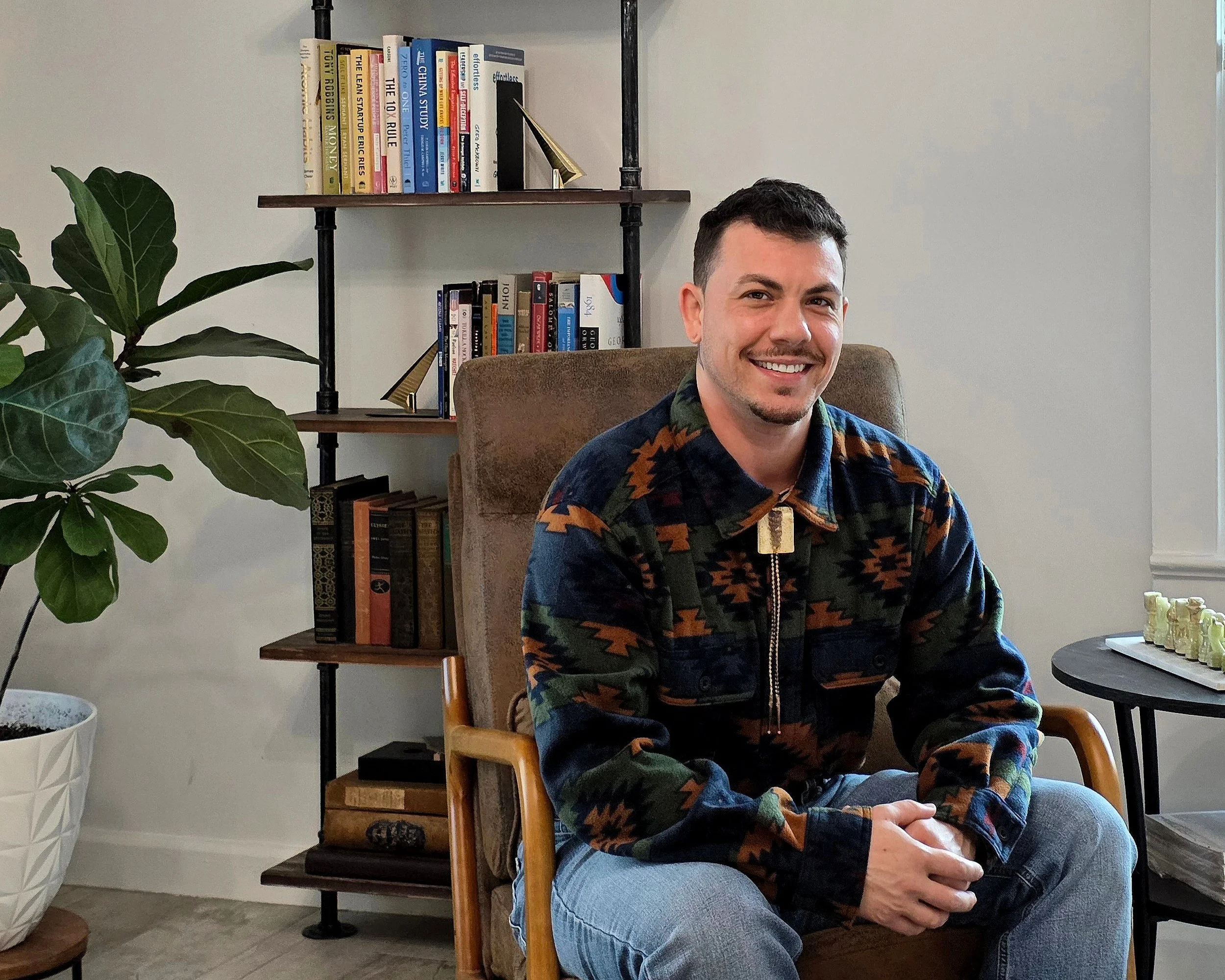 A man sitting in a brown armchair, smiling, wearing a colorful patterned jacket and jeans, in a room with a bookshelf, a large green plant, and a chessboard on a round table.