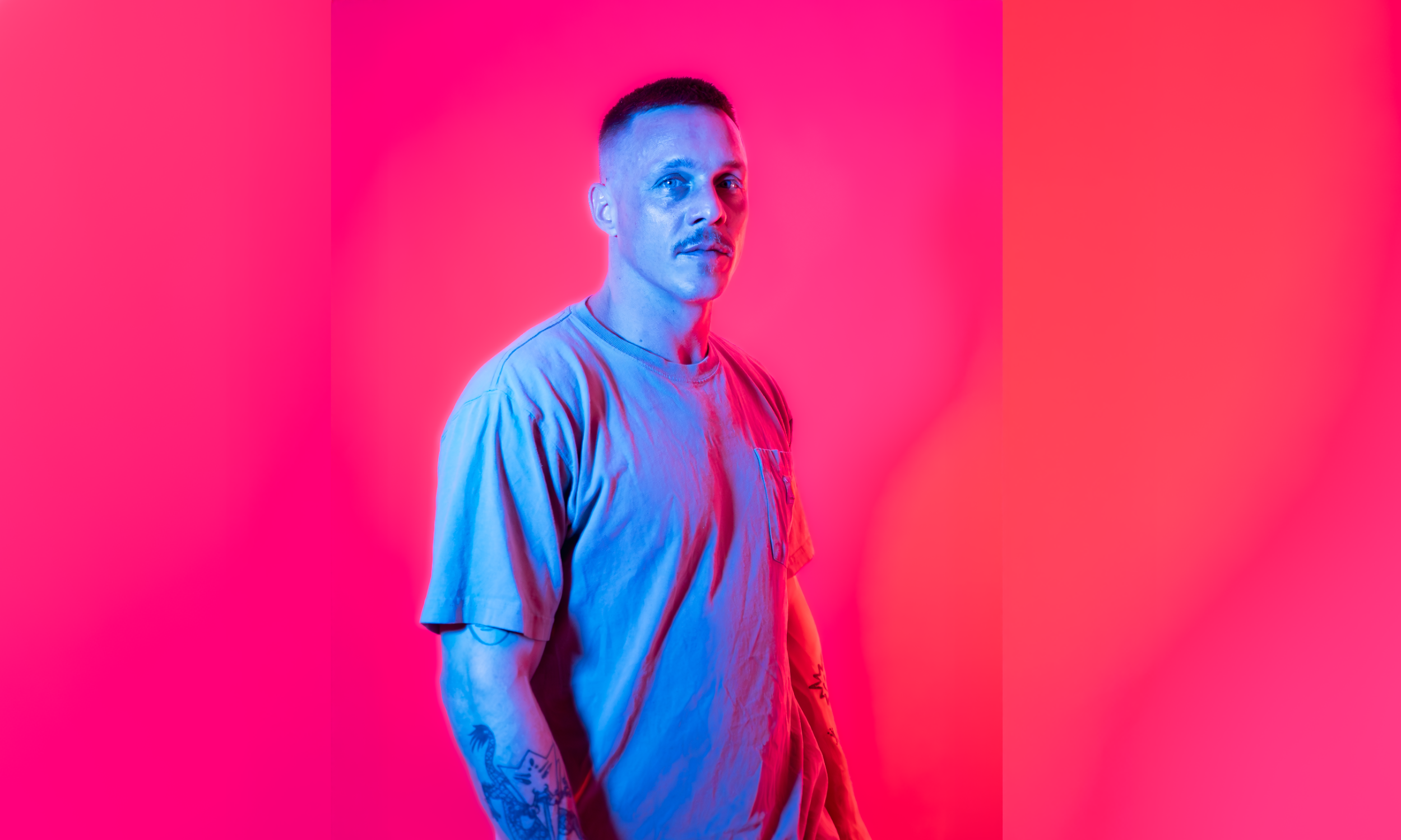 A man with short hair and tattoos on his arm wearing a loose shirt stands in front of a pink and red background