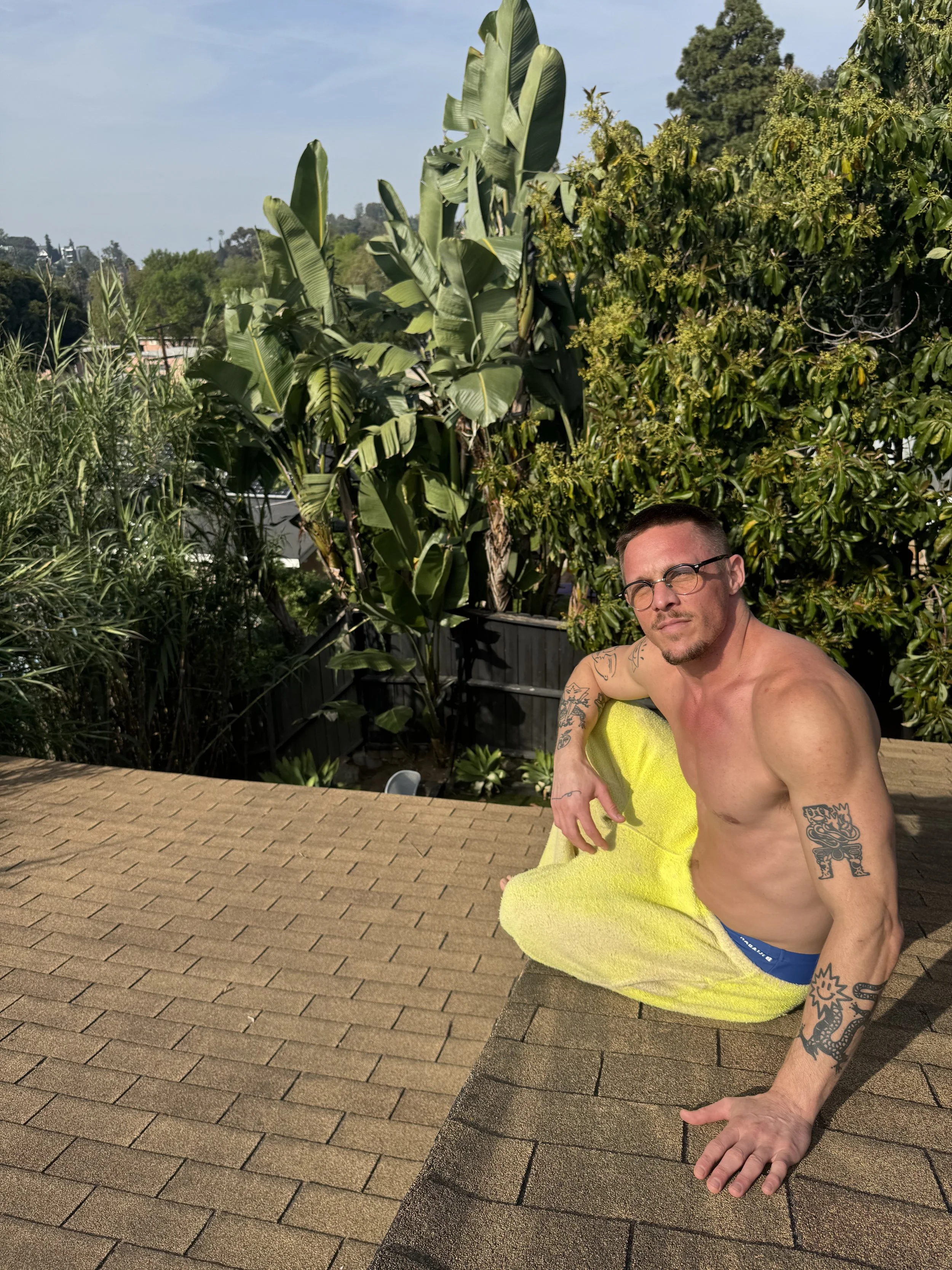 A shirtless man with tattoos on his arms and glasses sitting on a roof with a yellow towel wrapped around his waist, surrounded by lush green trees and a clear sky.