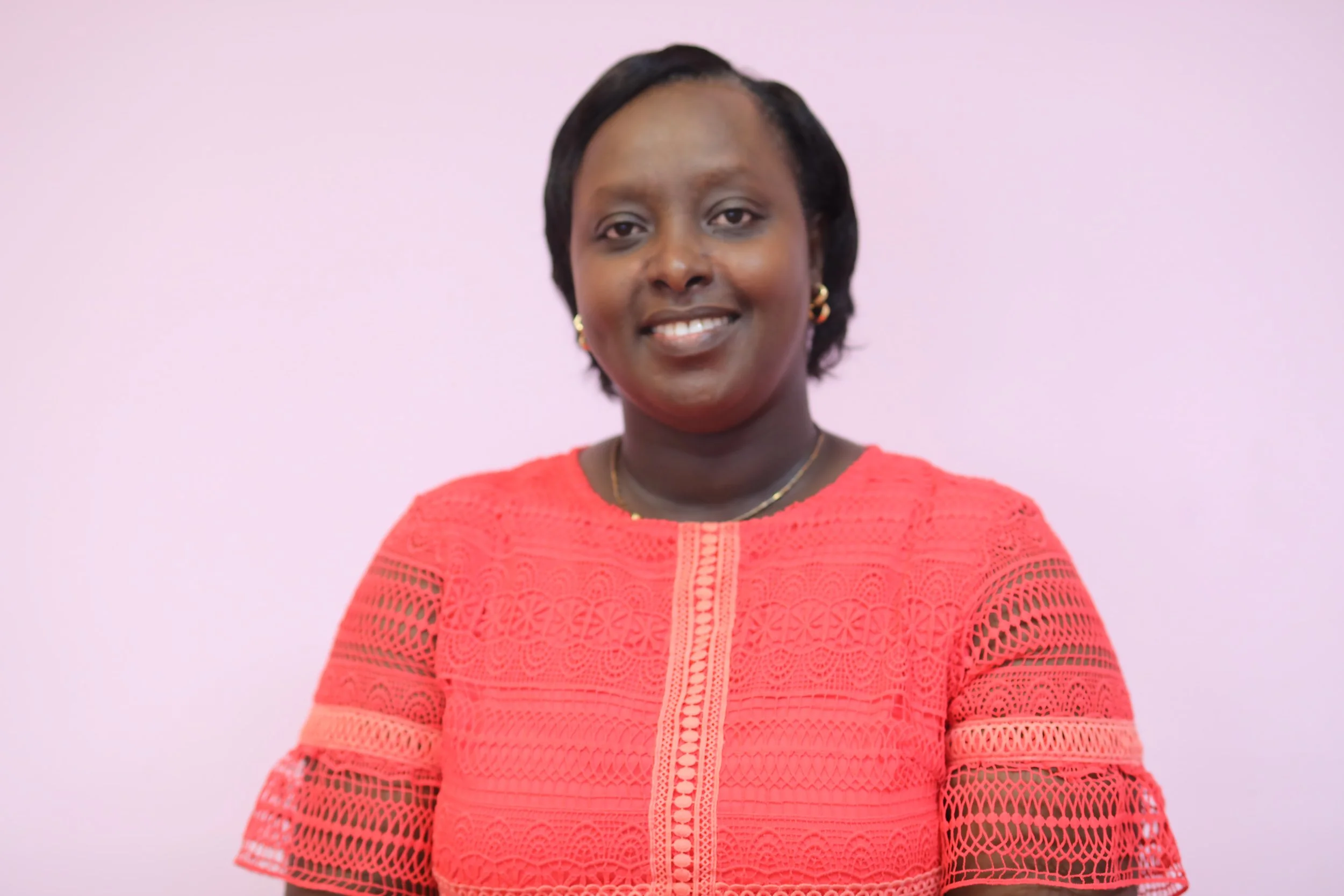 A woman with short black hair, smiling, wearing a red lace dress with intricate patterns, gold earrings, and a gold necklace, standing against a plain light pink background.