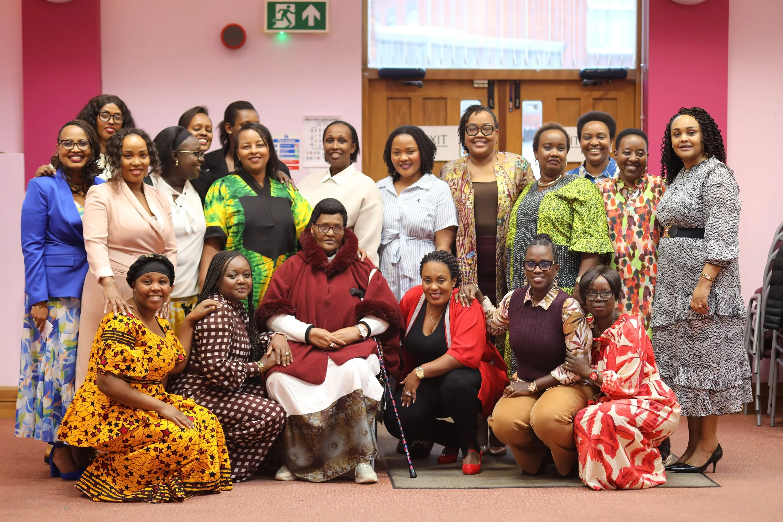 A group of women posing inside a building with pink walls, some wearing colorful traditional clothing and others in formal attire, standing and kneeling around an elderly woman with a cane, all smiling for the photo.