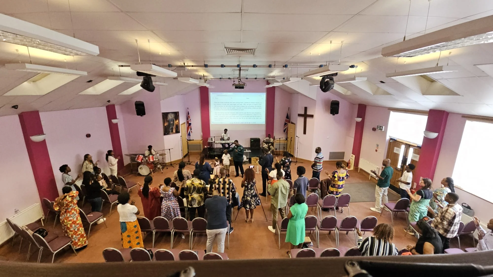 People gathered in a church or community hall with purple walls and a large cross. They are participating in a service or event, some singing or praying, with a band playing on stage, and a large screen displaying text behind them.