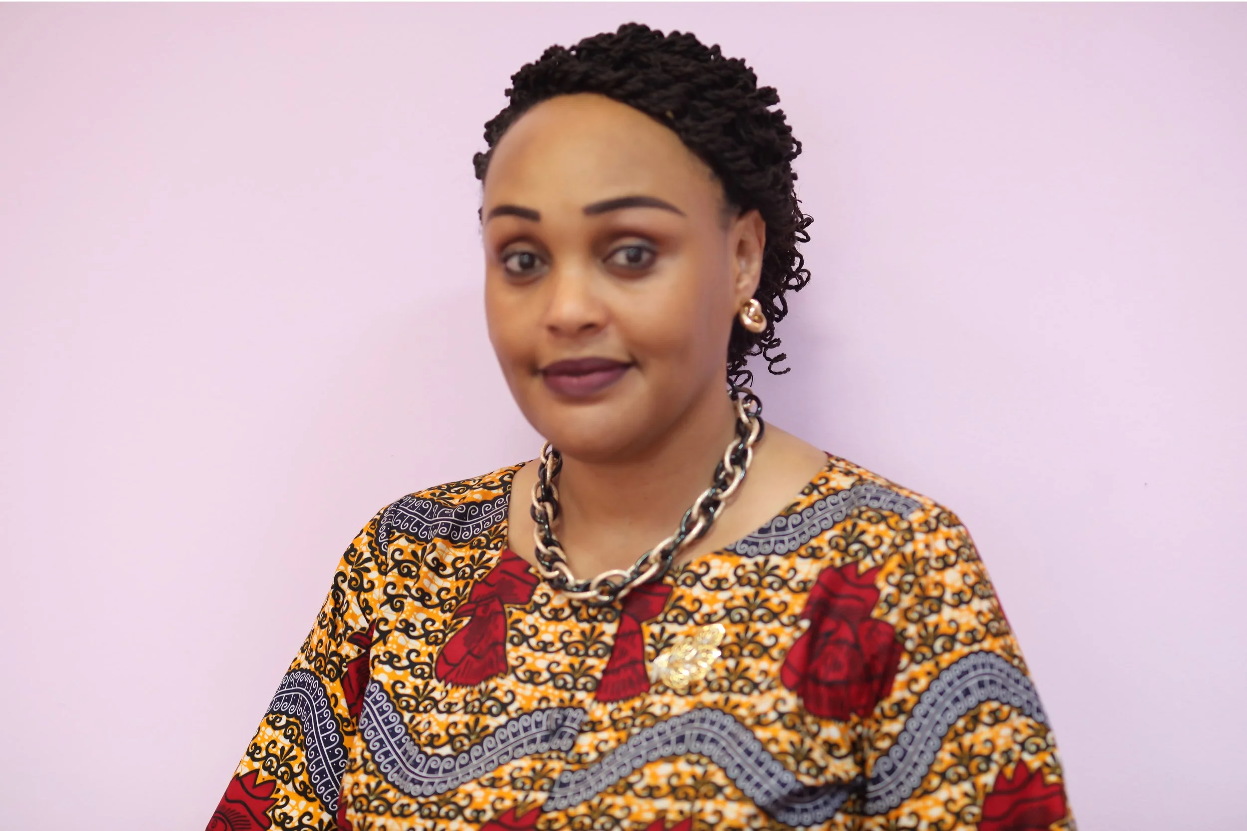 A woman with short curly hair, wearing a colorful patterned dress, gold jewelry, and a necklace, standing against a light purple background.