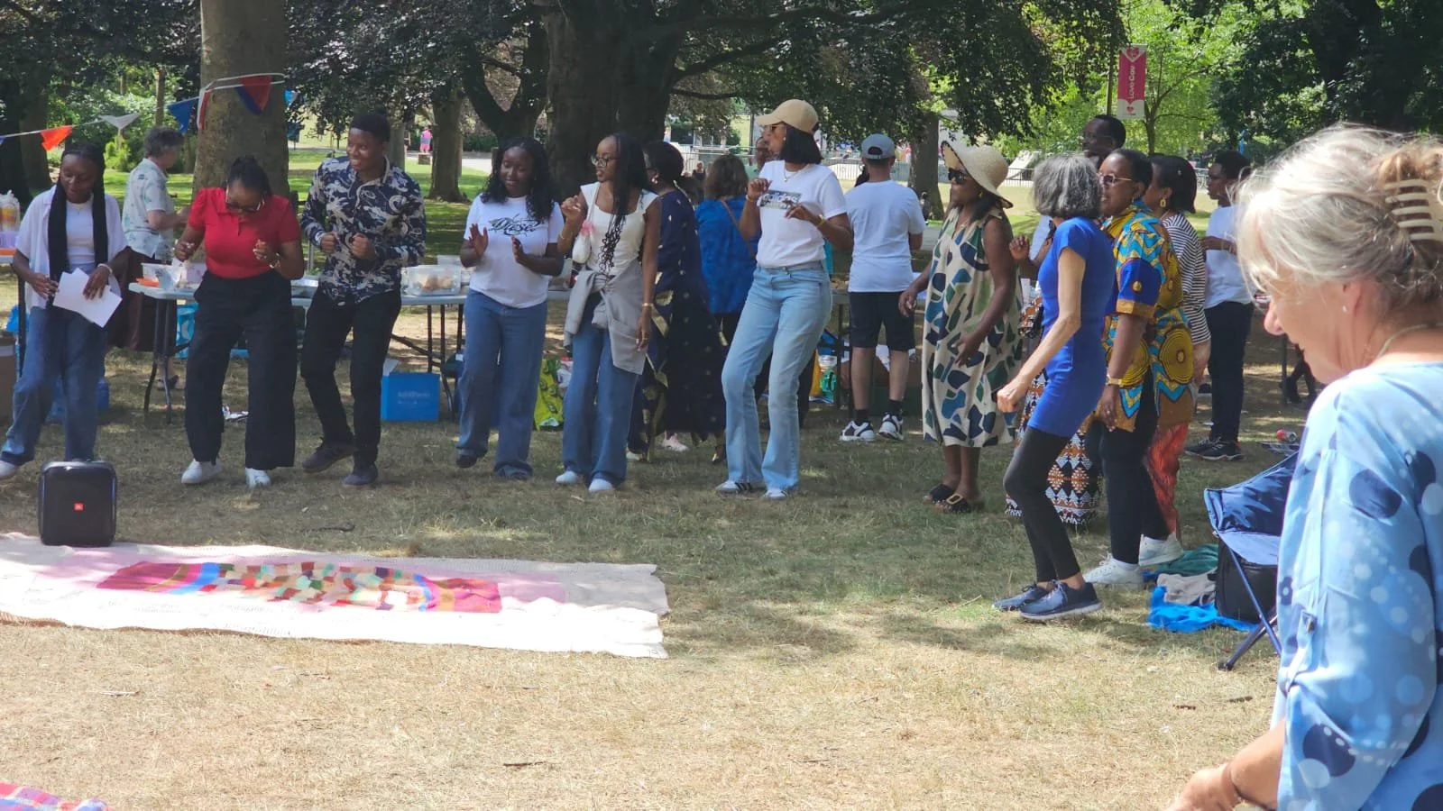 A group of people dancing outdoors in a park, some wearing colorful clothing, under large trees with banners and tables in the background.