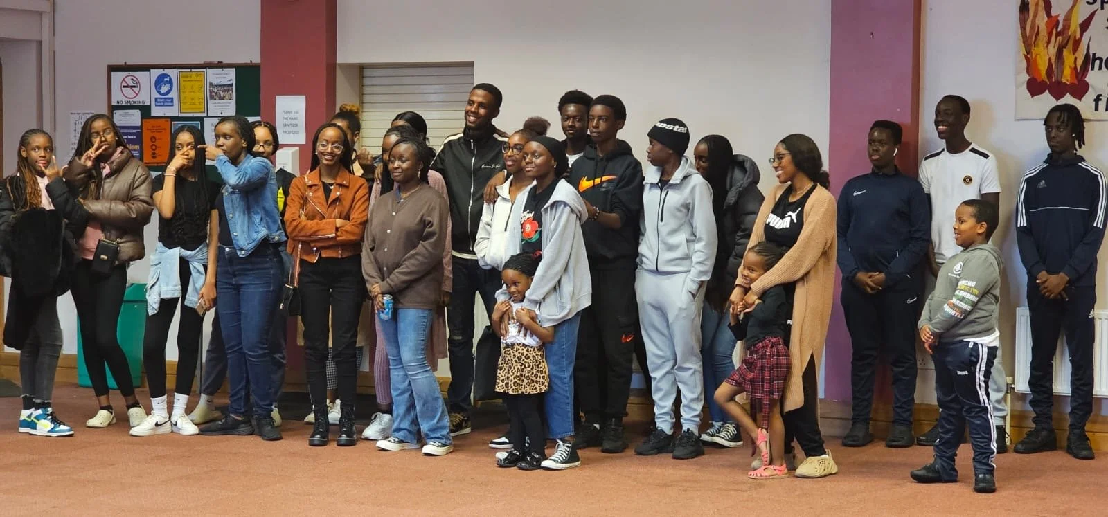 A group of diverse people, including children and adults, gathered indoors, some smiling and chatting.