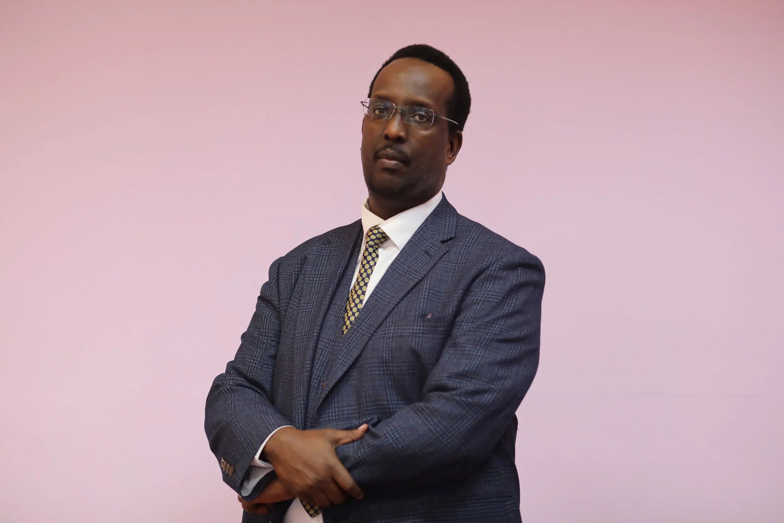 A man in a dark gray checkered suit, white shirt, and patterned yellow tie standing with arms crossed against a plain pink background.