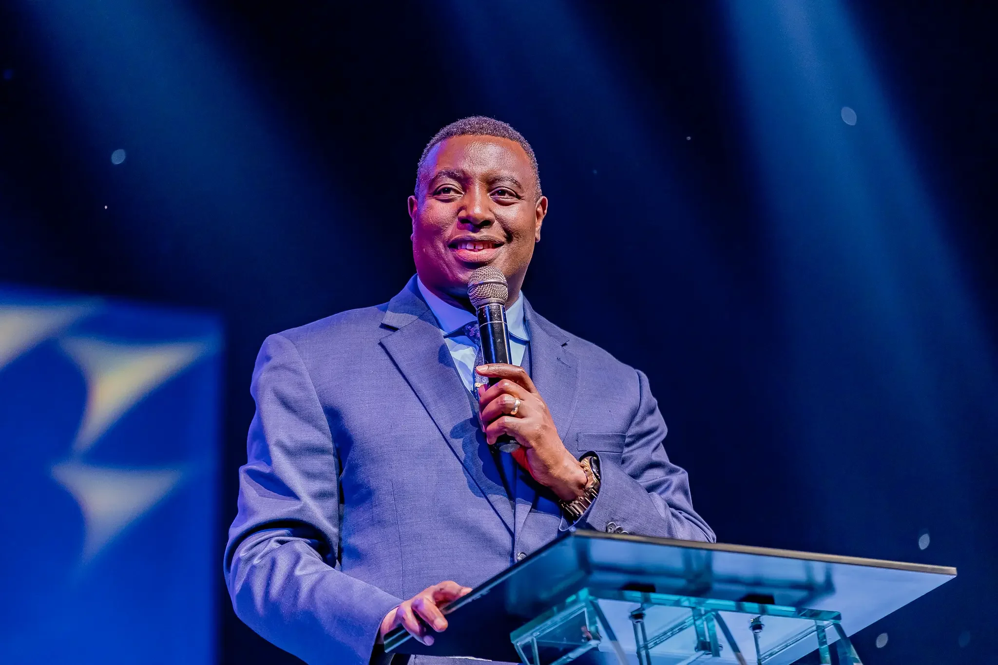 African American man in gray suit speaking into a microphone at a podium on a stage with blue lighting.
