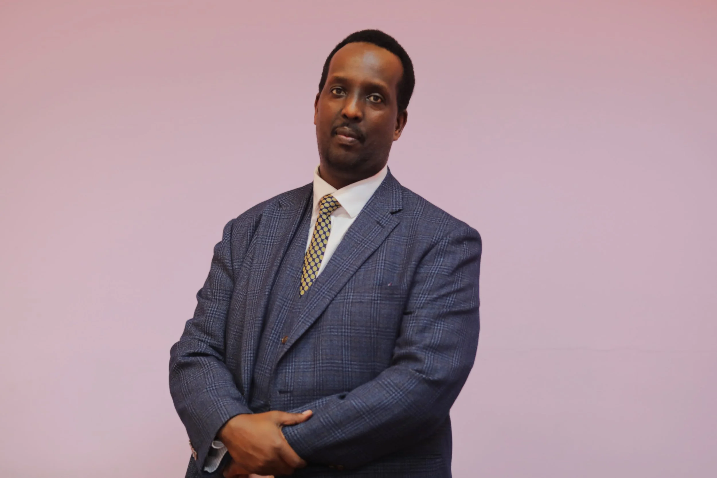 Man in a dark blue plaid suit with a white shirt and yellow patterned tie, standing against a light pink background with arms crossed.