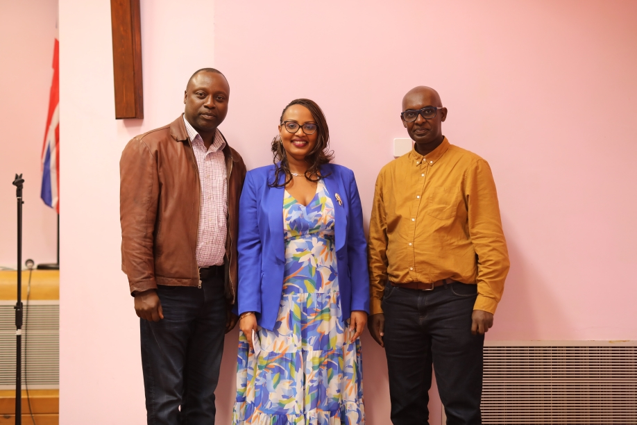 Three people, two men and one woman, standing in a room with a pink wall. The woman is in the middle, wearing glasses, a blue blazer, and a patterned dress. The man on the left is wearing a brown jacket and dark jeans. The man on the right is wearing a mustard-colored shirt and black pants.