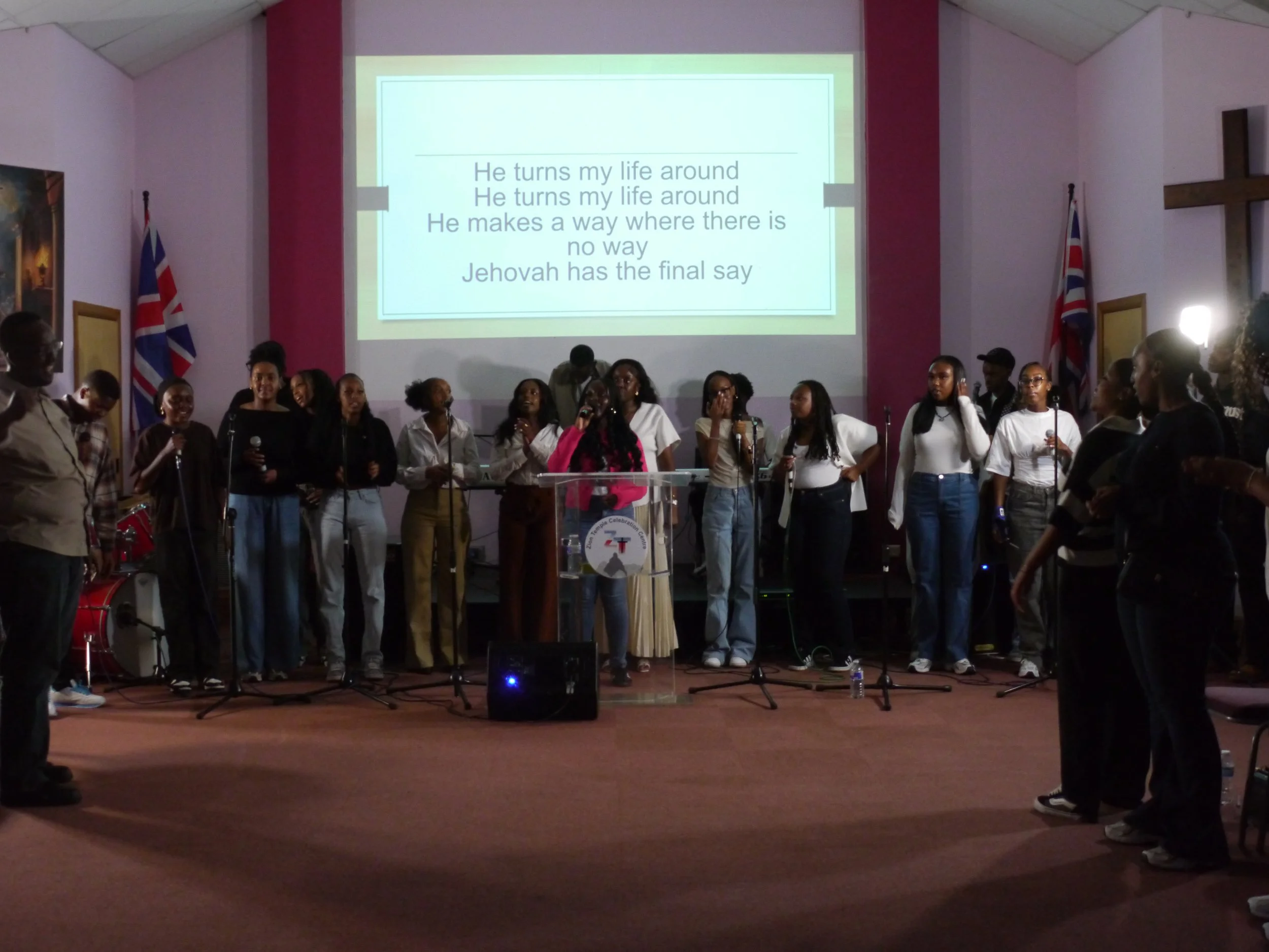 A group of people standing on stage in a church, singing or performing, with lyrics projected on a screen behind them. The words on the screen are part of a song about Jehovah. There are two flags on either side of the stage, and a cross on the wall to the right.