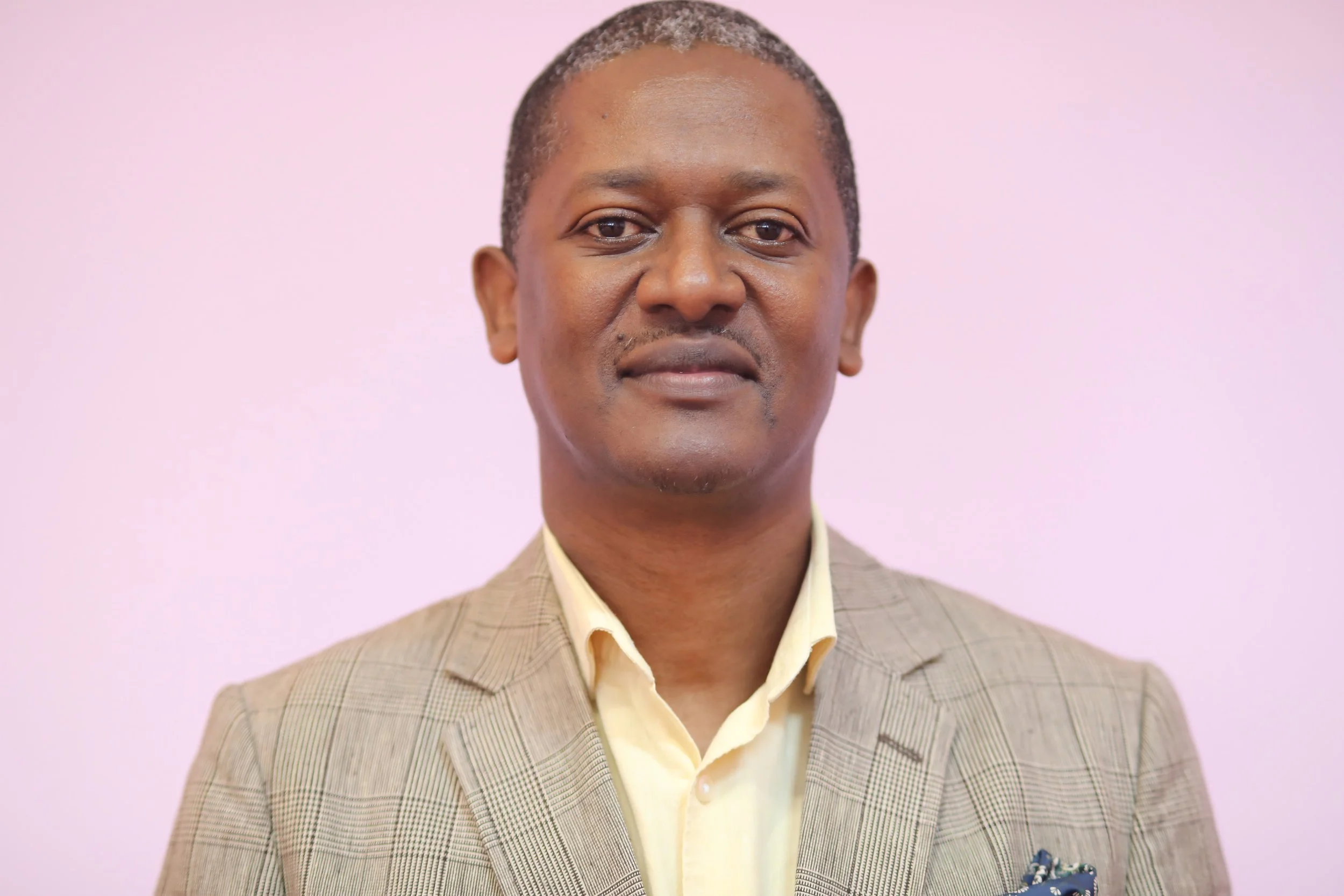 A portrait of a middle-aged man with short, dark gray hair, wearing a beige plaid blazer and a yellow collared shirt, standing against a plain light pink background.