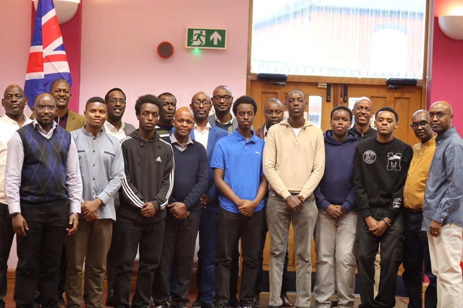 Group of 16 men standing together in a room with pink walls, a UK flag, and a wooden door, posing for a photo.