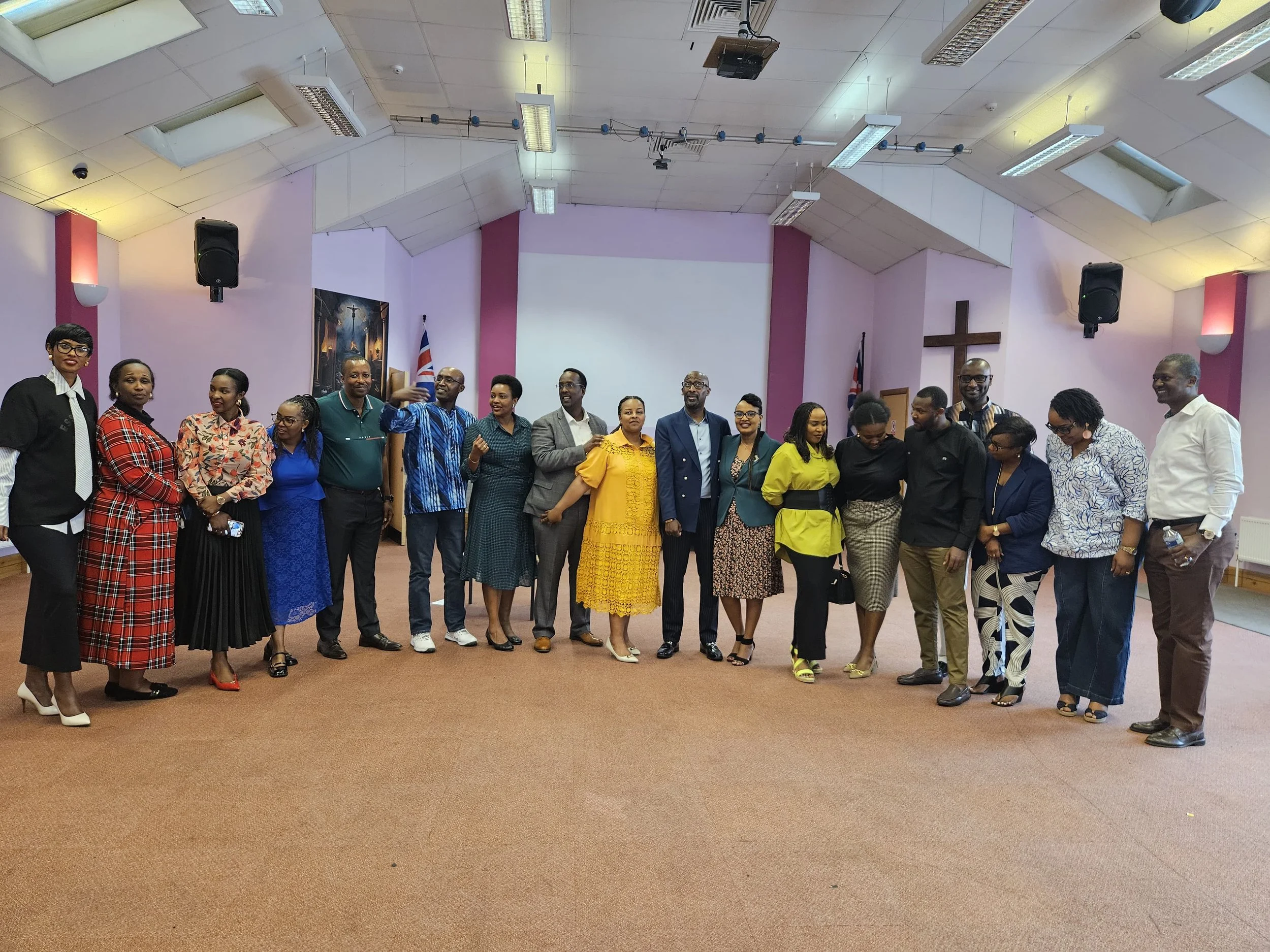 Group of people standing inside a church or community hall, posing for a photo. The group includes men and women dressed in formal and semi-formal clothing, with flags and religious symbols visible in the background.