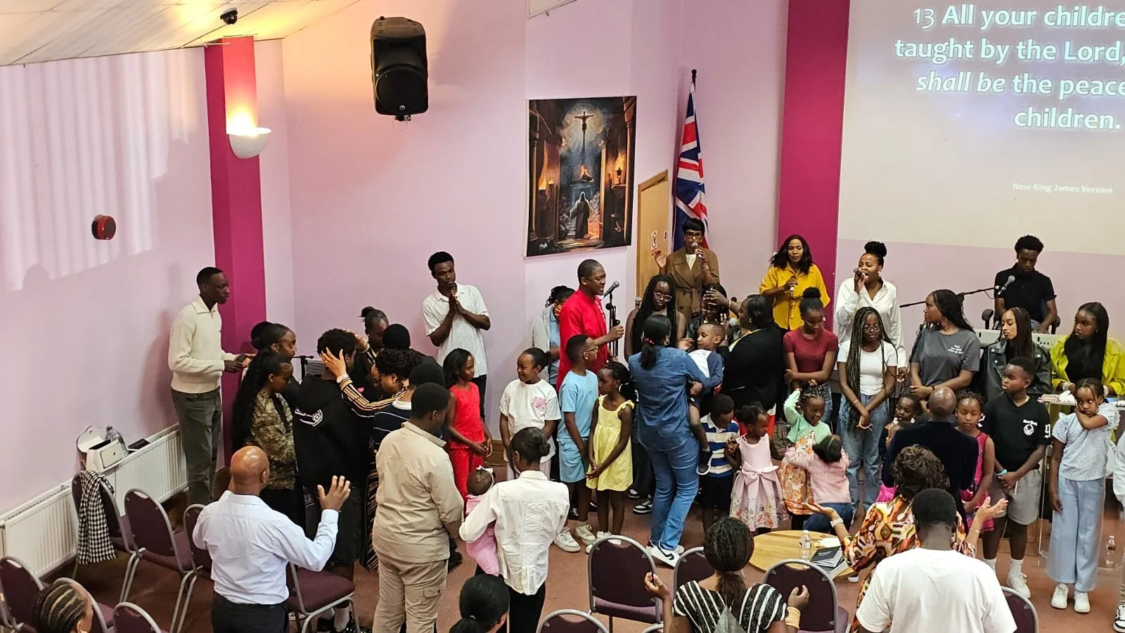A diverse group of people, including children and adults, gathered in a church for a service or event. Some people are standing near the front, while others are seated or standing around. The background features a large screen with religious text, a painting, a Union Jack flag, and pink and purple walls.