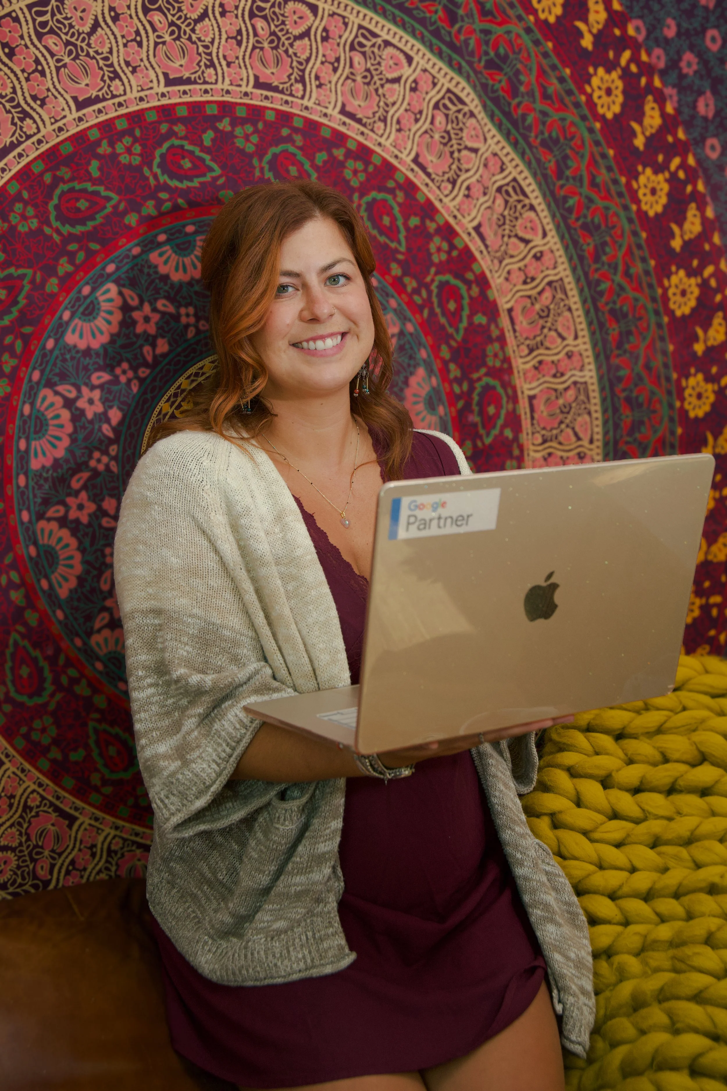 A woman with reddish-brown hair smiling while holding a silver laptop with a "Google Partner" sticker, sitting against a colorful mandala tapestry background.